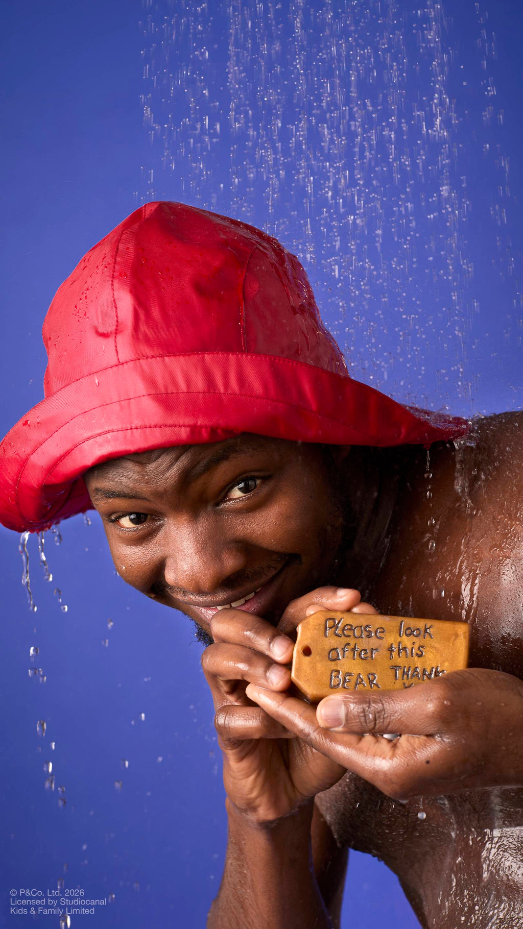 A model wearing a red rain hat stands under running shower water. They hold a brown, luggage-tag-shaped soap featuring the embossed message ‘Please look after this bear, thank you’. Droplets cascade down against a solid blue background.