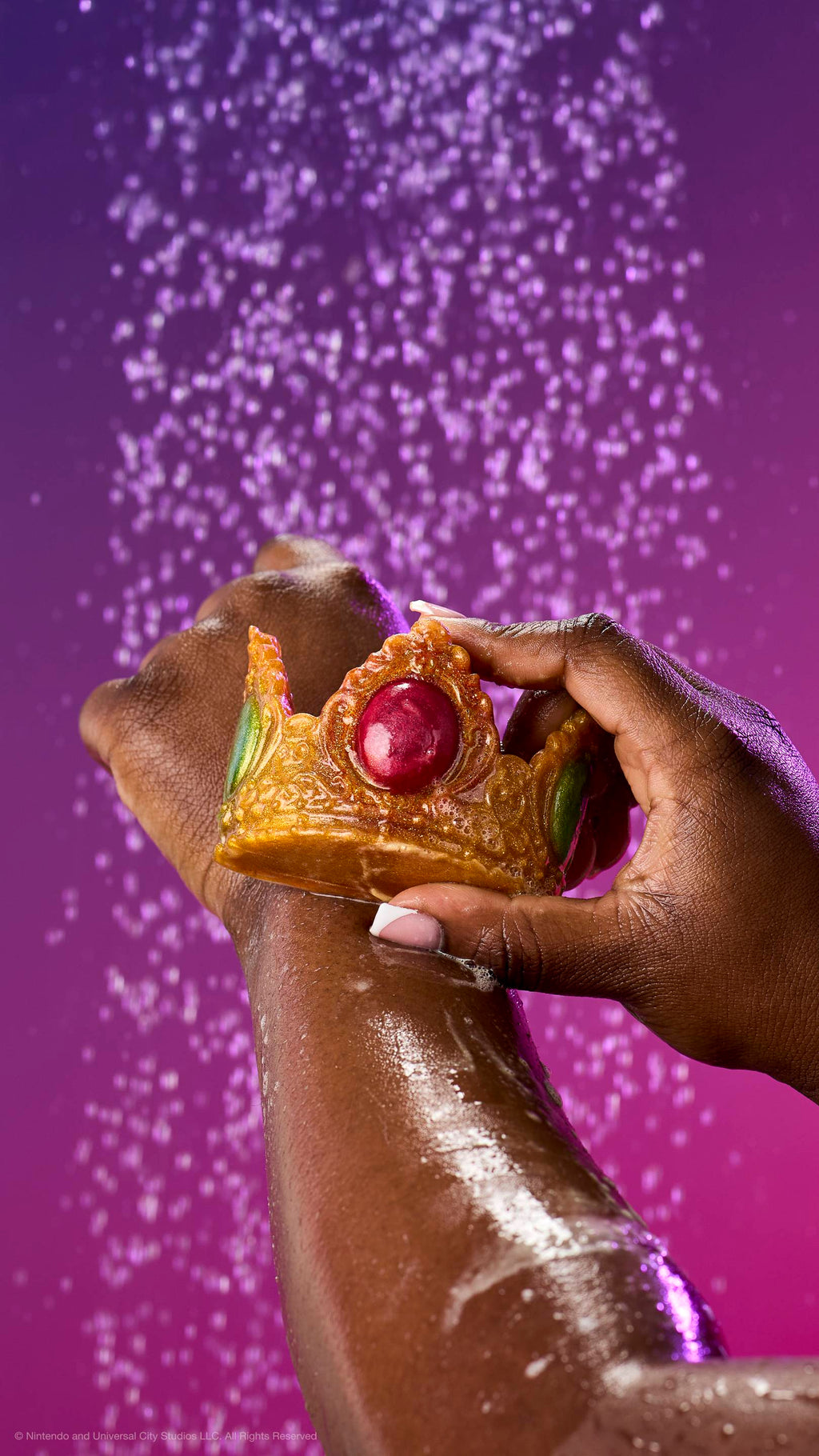 A hand glides the shimmering gold Princess Peach crown soap over a soapy forearm under a shower spray. Features red and green jewels against a purple-pink gradient background.