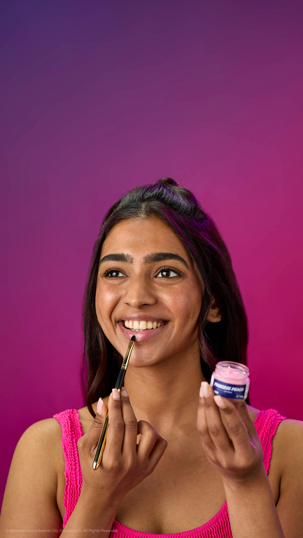 A woman in a pink top smiles while applying pink Princess Peach lip jelly with a gold brush. She holds the open glass pot against a purple-magenta gradient background.