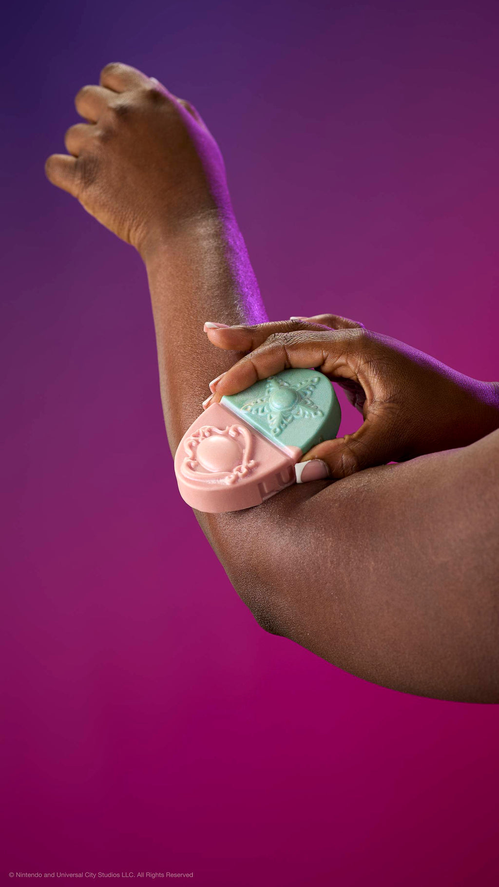 A hand glides the pink and teal Princess Power body balm over an elbow. Features embossed brooch and star designs. Against a purple-magenta gradient with plastic-free glitter.