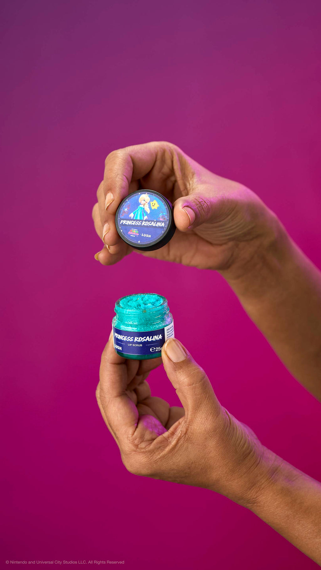 Two hands hold an open pot of turquoise Princess Rosalina lip scrub and its lid against a purple background. The lid features Princess Rosalina and a Luma character.