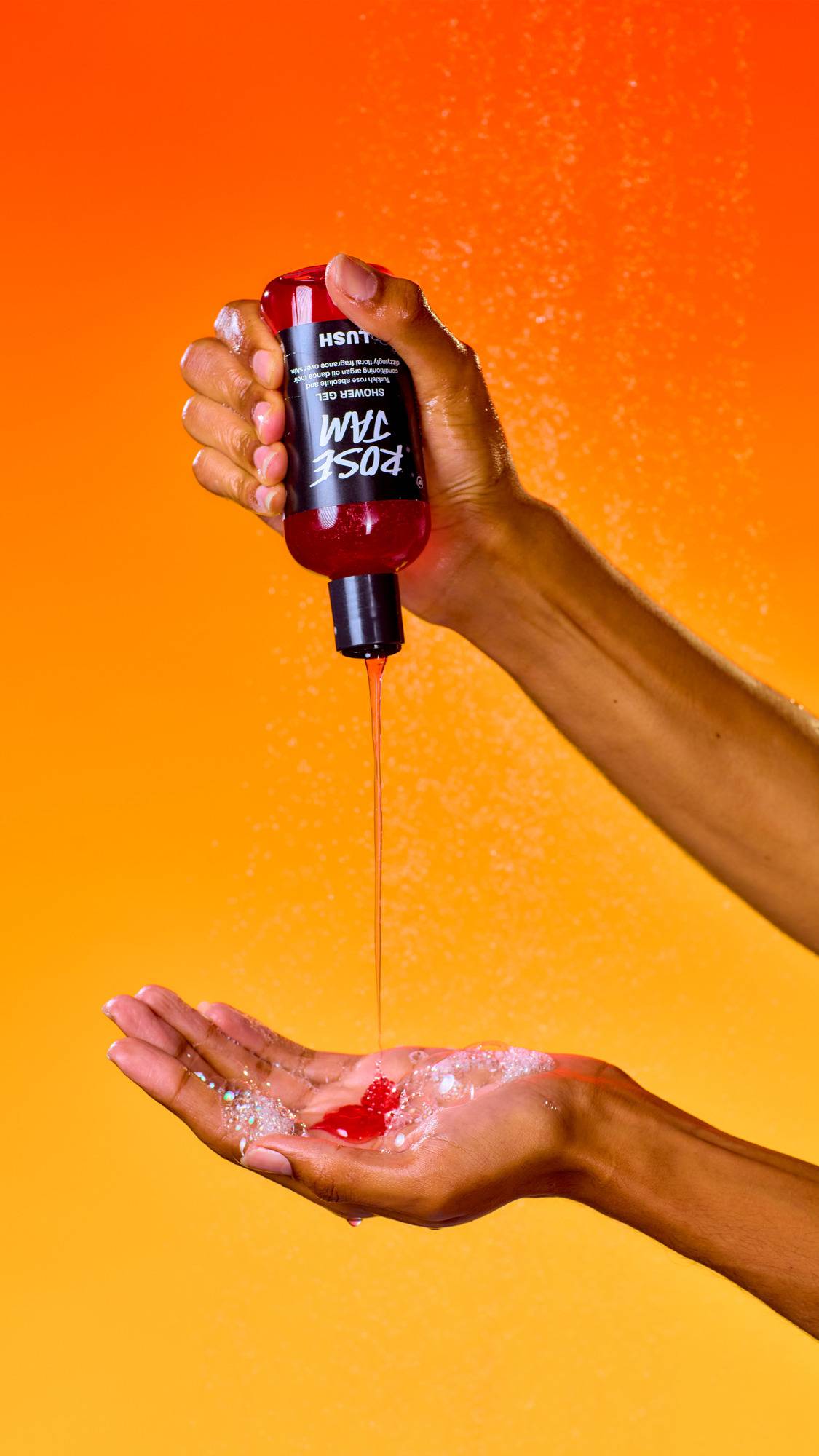 An upside-down bottle of red Lush Rose Jam shower gel is squeezed, pouring a steady stream of red liquid into a waiting, soapy palm. A fine water mist spray fills the air against a vibrant, solid orange background.