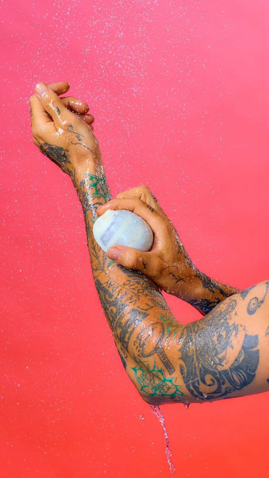 Tattooed arms and hands hold a smooth, light-coloured Scrubee bar under a spray of water against a vibrant pink background. Water droplets cover the glistening skin and stream down from the elbow.