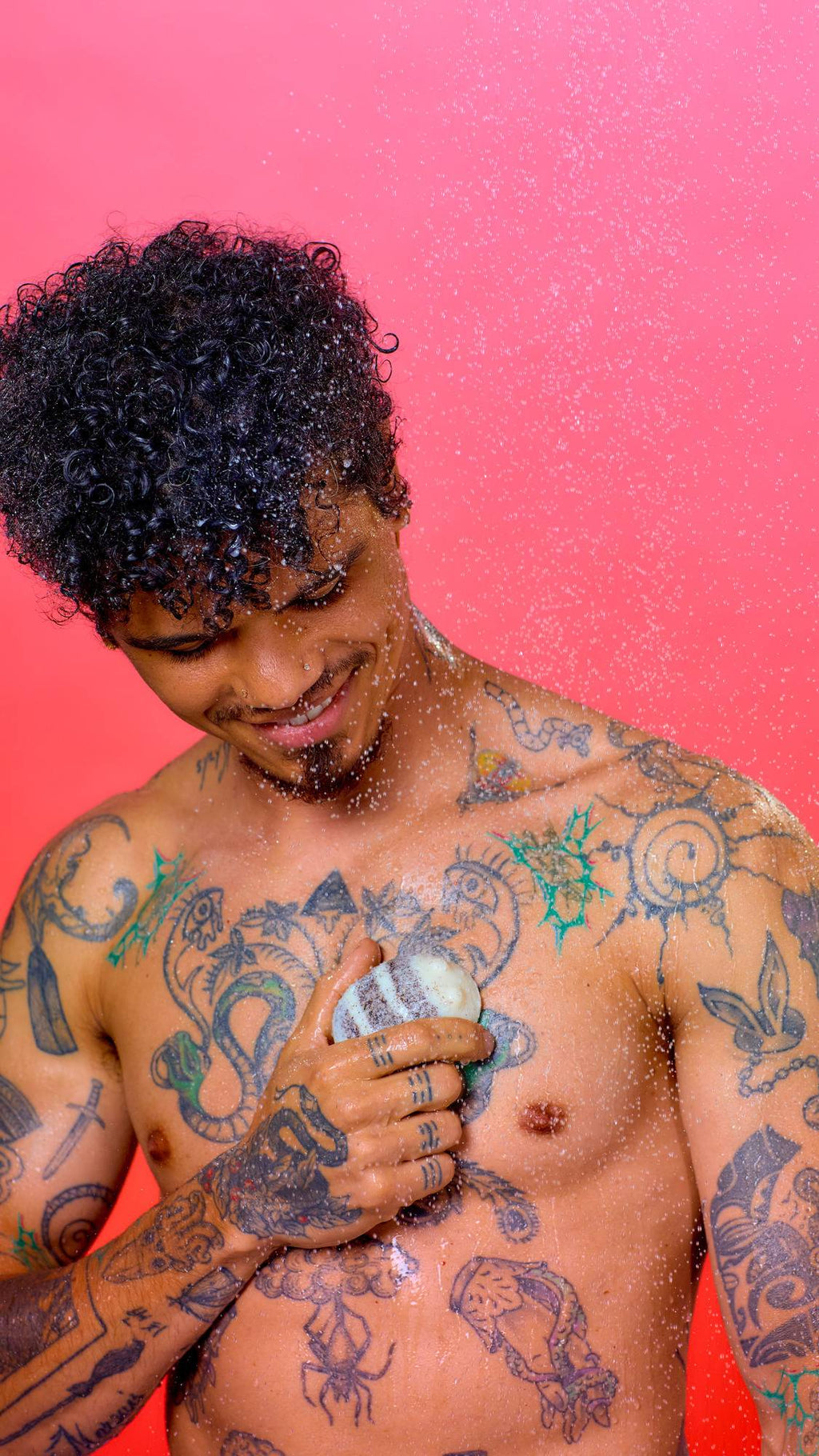 A model with curly black hair and numerous colourful tattoos stands under a spray of water against a pink background. They hold a striped, bee-shaped solid bar against their chest as water droplets splash over them.