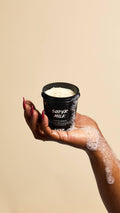 A hand with long red fingernails holds an open pot of "Super Milk" Protein Shampoo. Soapy bubbles run down the side of the black container and onto the hand, highlighting the creamy texture of the product inside against a beige background.