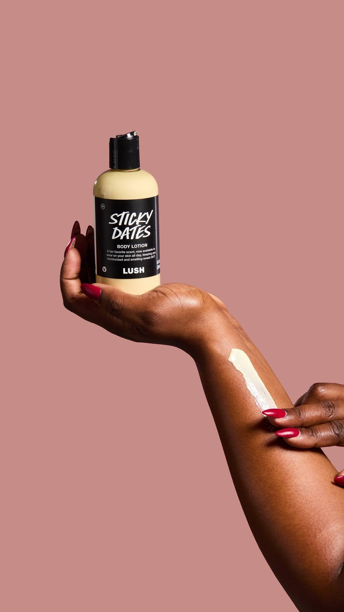 A hand with long, deep red nails massages a streak of creamy, sticky dates lotion into the skin of a forearm against a dusty pink background.