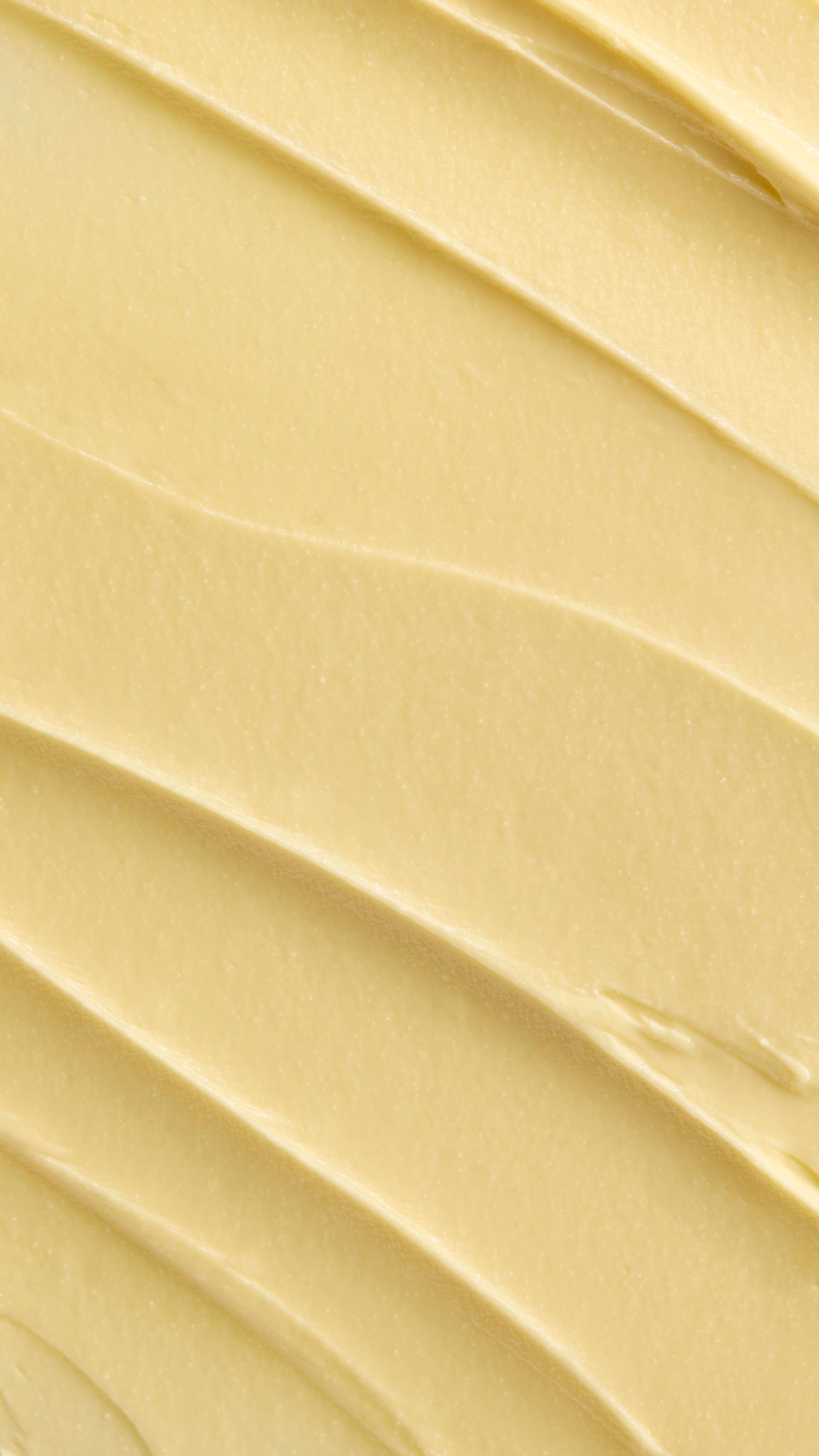 A super close-up image showing diagonal, overlapping smears of the thick, creamy, pale yellow Lush "Acai & Sacha Inchi" Vitamin E facial cleanser. The smooth product has a matte finish and subtle ridged textures.