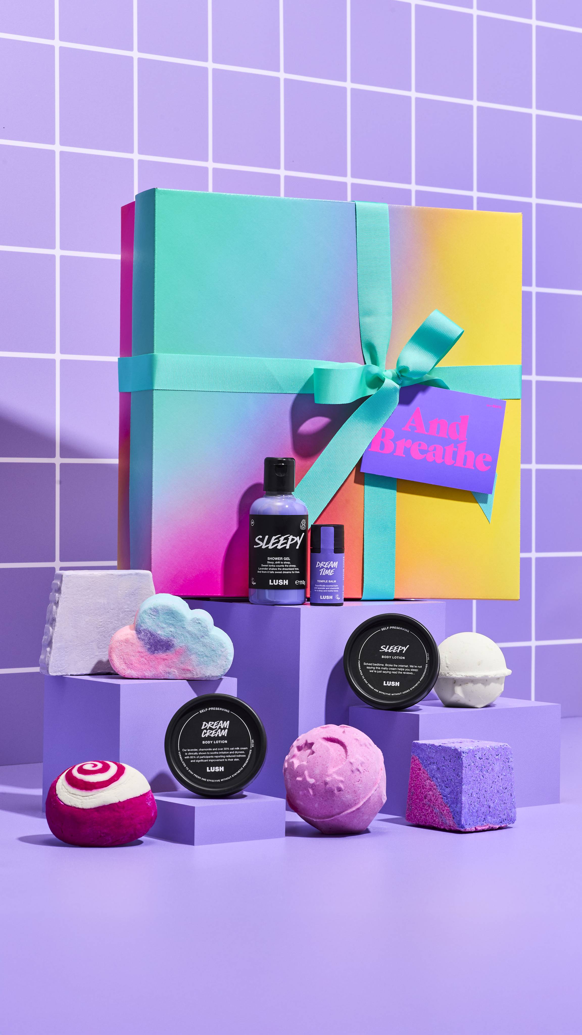 The image shows the colourful And Breathe Lush gift box sitting on a pastel purple platform on a purple-tiled background. The ten included Lush products surround the box on separate smaller platforms.
