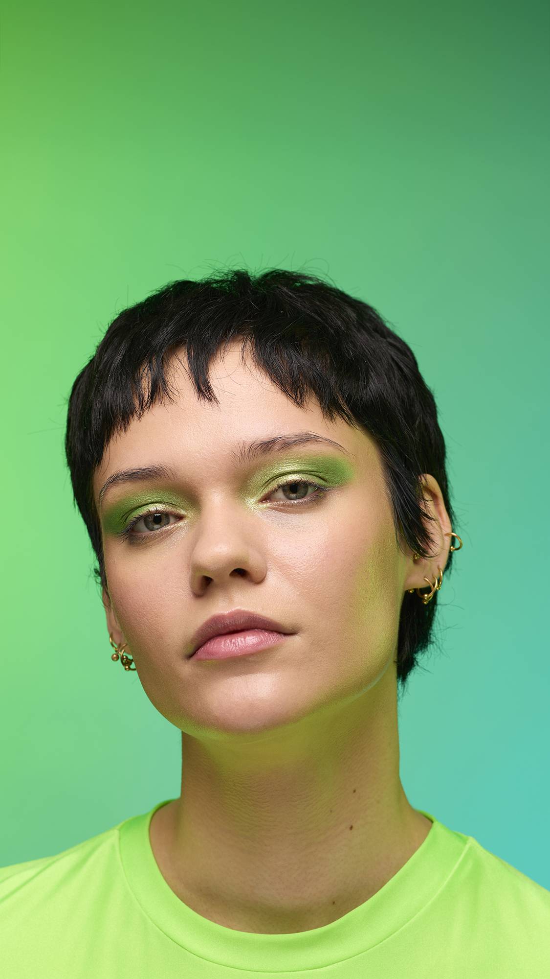 The image shows a close-up of the model's face on a pale blue-to-green gradient background. The model's make-up is done with the focal point being a subtle, jade-green, shimmering eyeshadow made by the Aurora Glitter Crumble.
