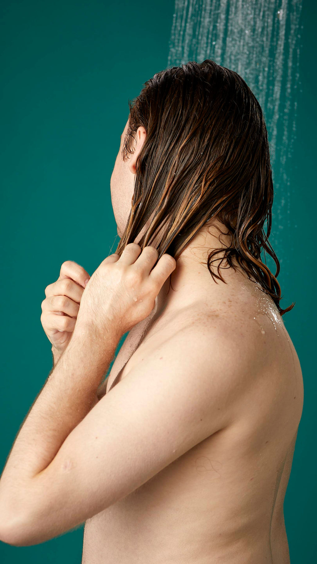 The image shows the model from the side while facing away, under running shower water. Their hair is wet and has been shampooed already. They are now running the Banana conditioner through the lengths and ends of their hair.