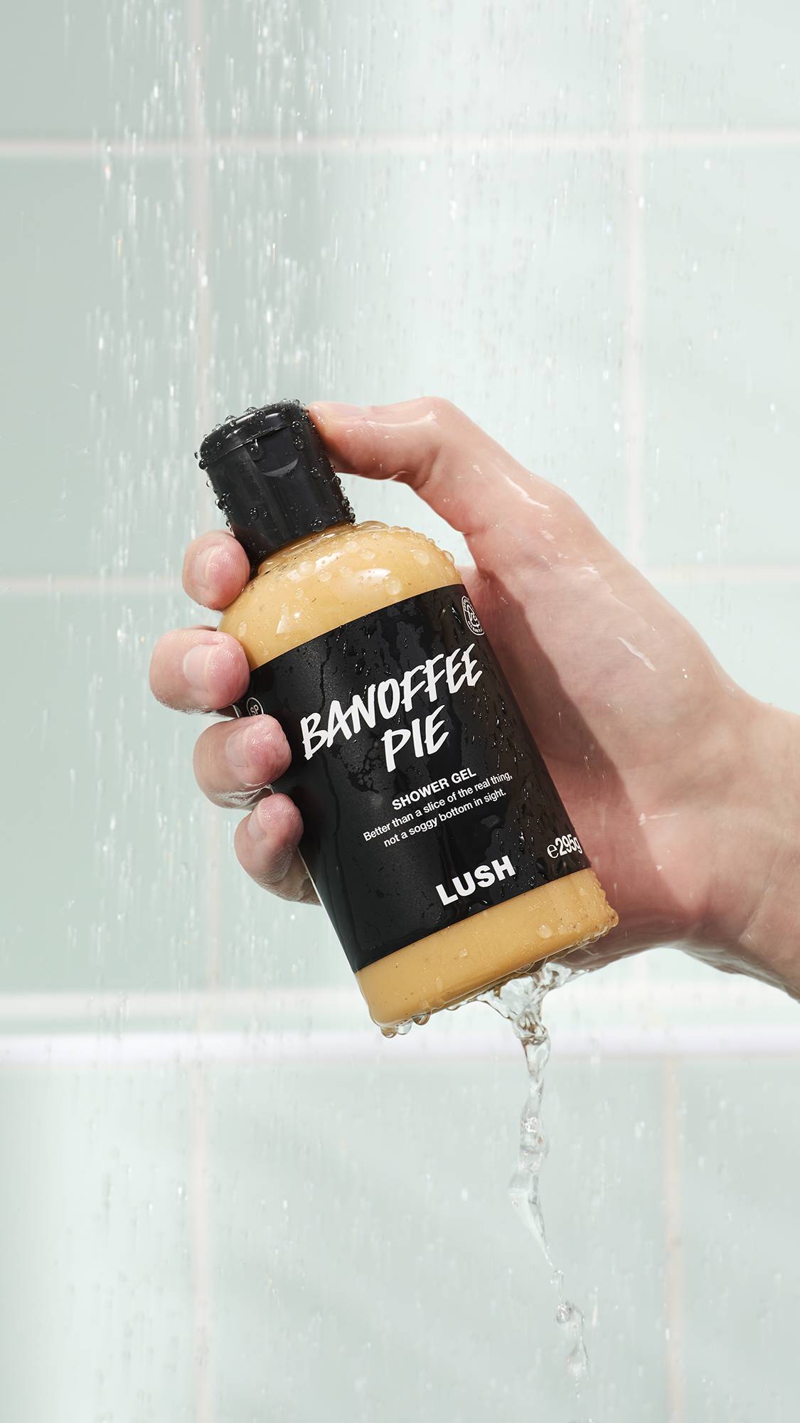 A hand holds a bottle of Lush Banoffee Pie shower gel under a stream of water in a shower. The clear bottle reveals the creamy yellow gel inside, and the black label features white text. Mint green tiles are in the background.