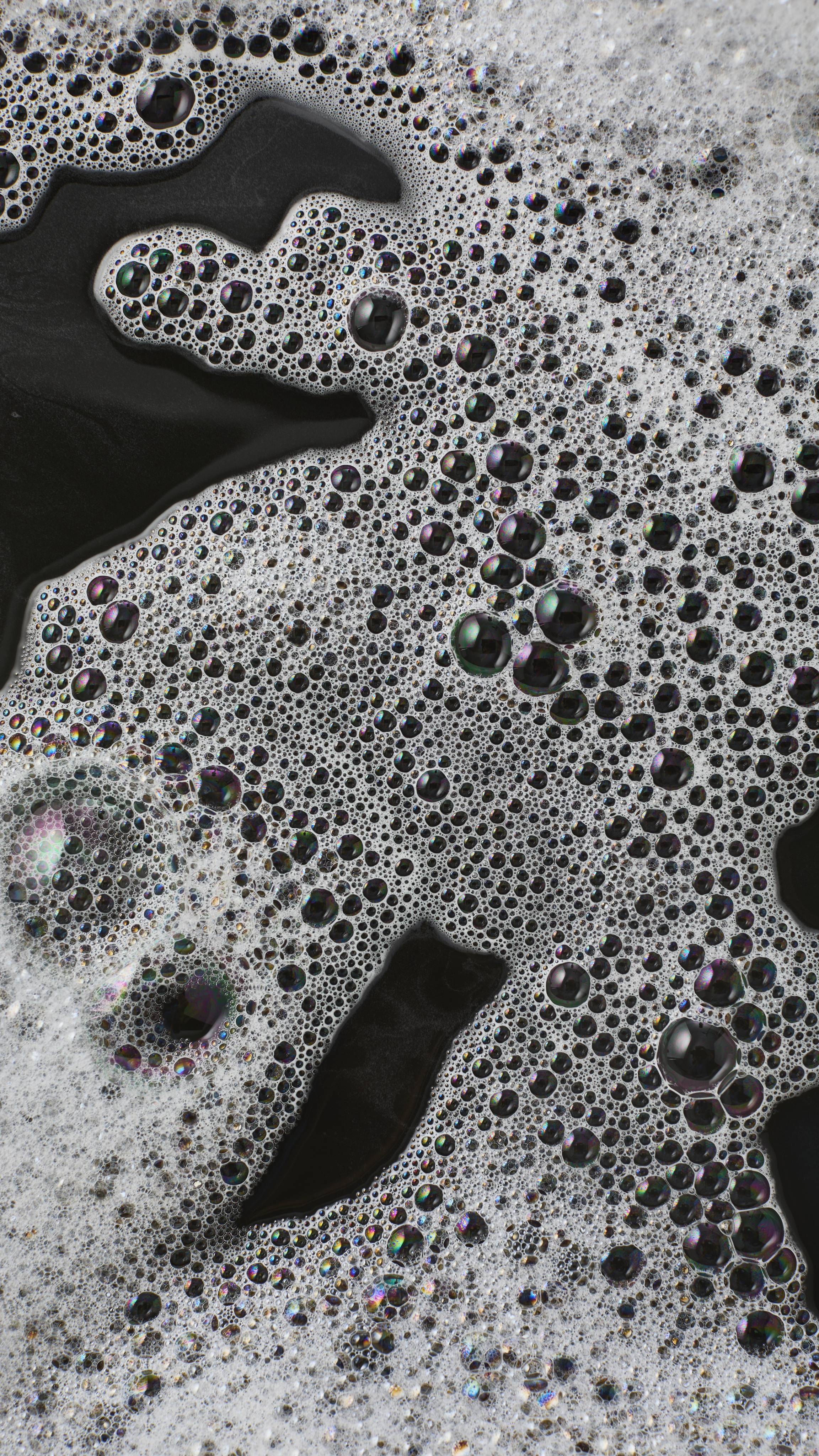 An abstract macro shot of thick white foam floating on dark, near-black bath water. The frothy surface is densely packed with a mixture of tiny white bubbles and larger, translucent bubbles reflecting iridescent colours.