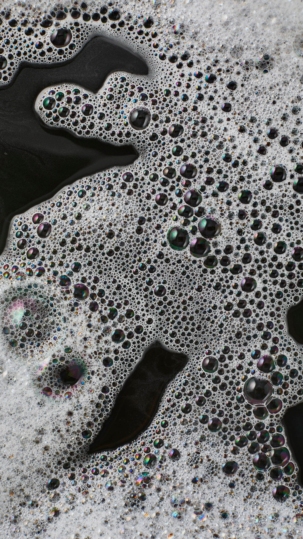 An abstract macro shot of thick white foam floating on dark, near-black bath water. The frothy surface is densely packed with a mixture of tiny white bubbles and larger, translucent bubbles reflecting iridescent colours.