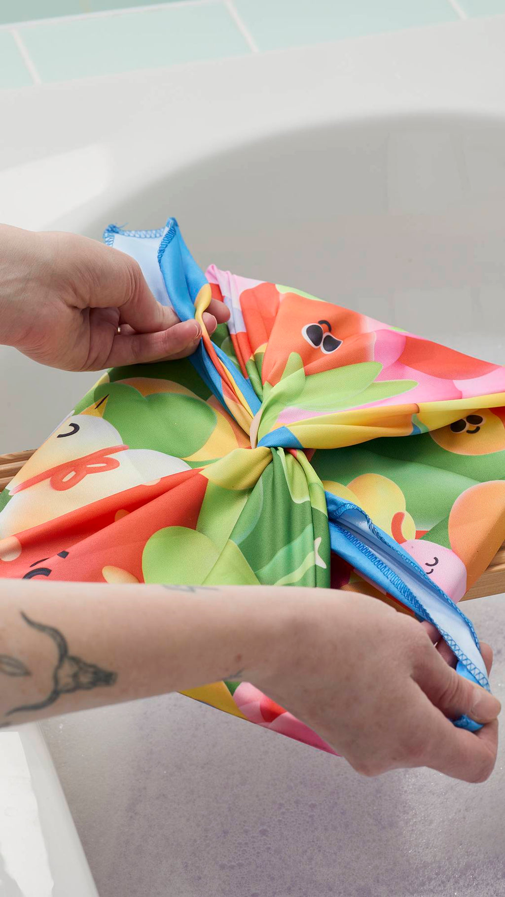 A model's hands are shown tying a "Blooming Lovely" Knot Wrap into a bow over a gift. The fabric features a colourful, 3D-style garden print. The gift rests on a wooden bath tray over a soapy bathtub in a bright bathroom.
