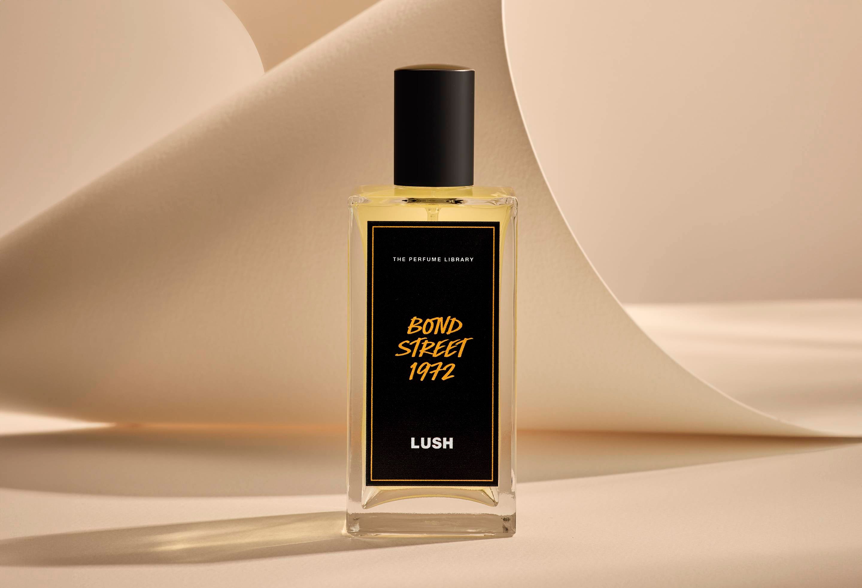 The image shows the Bond Street 1972 glass perfume bottle. It is sitting on a soft, beige background with curved, cream paper creating soft, gliding shapes behind the product.