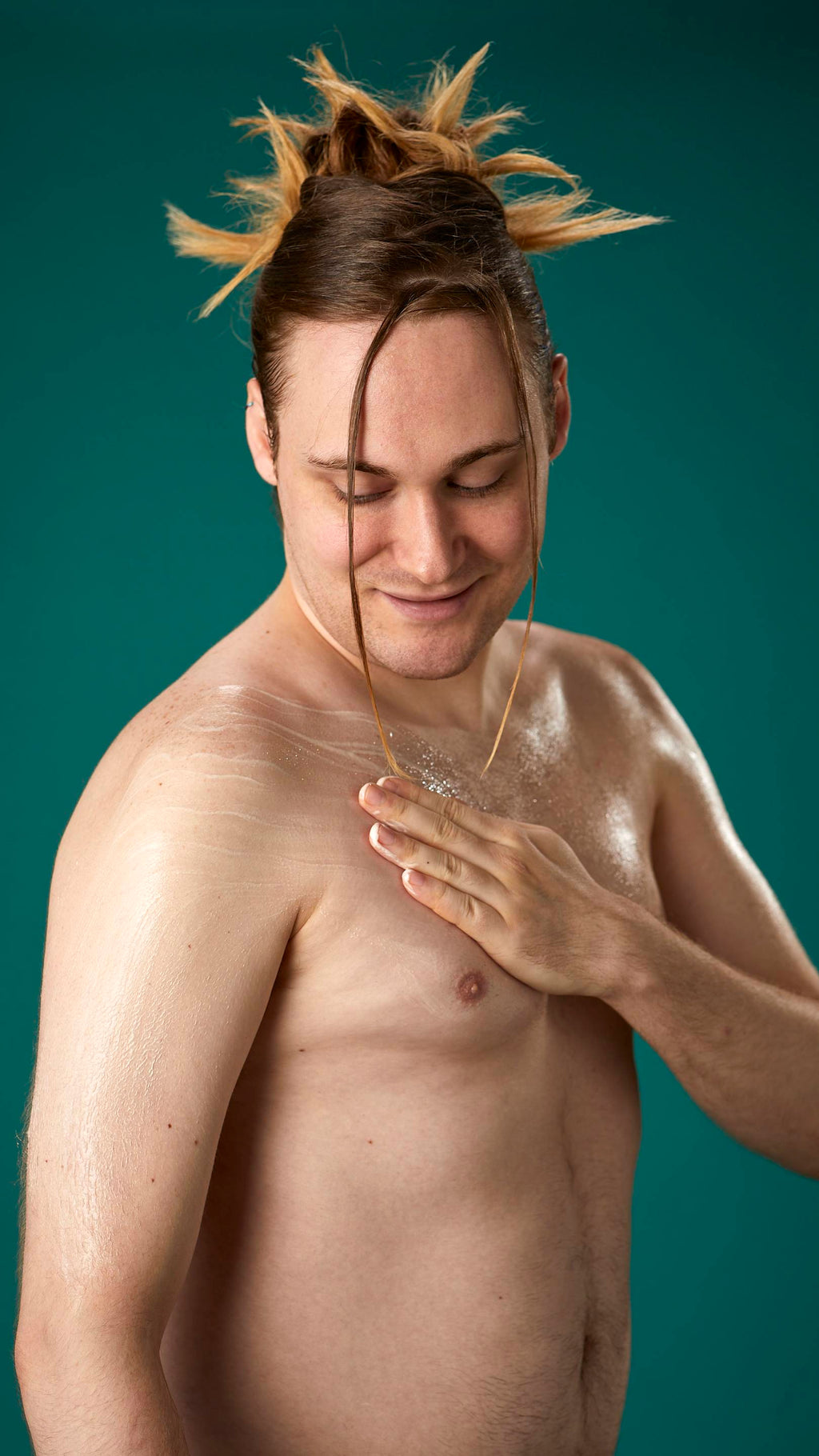 The image shows a topless model on a teal background. They are gently soothing the Braziliant body butter in circular motions over their chest and upper arm.