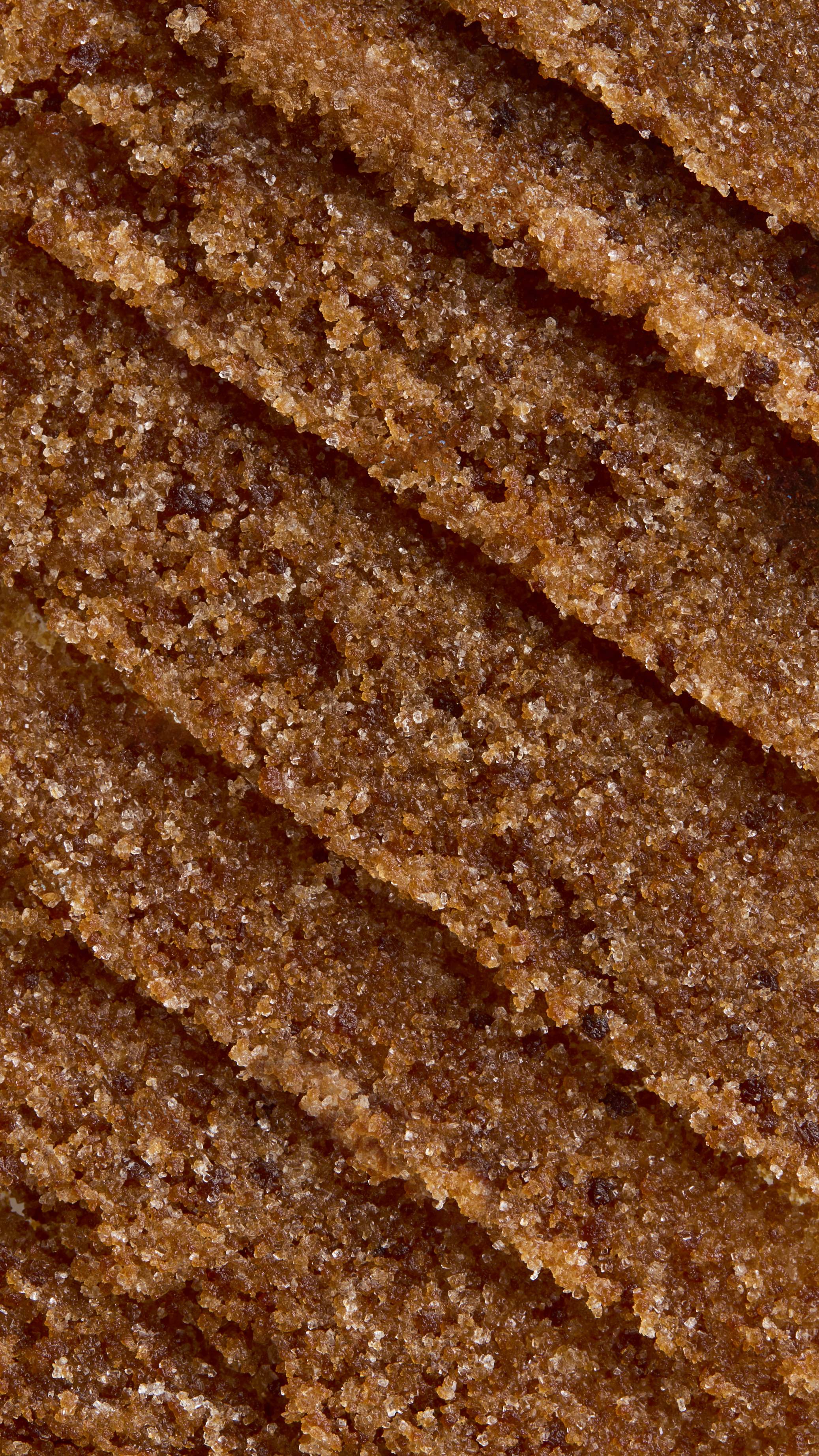 A close-up, aerial view of textured, thick caramel-coloured Buzz lip scrub. The texture is rich and gritty, arranged in several diagonal, overlapping swoops that create ridges and valleys.