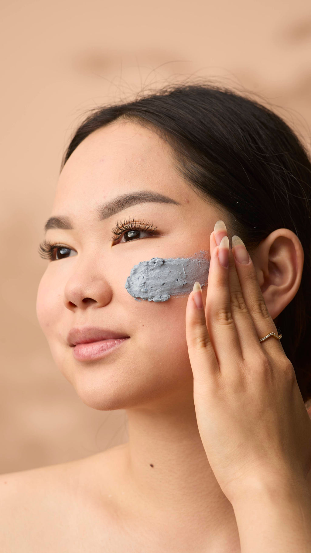 A close-up portrait of a woman applying Catastrophe Cosmetic face mask to her cheek. She uses her fingers to spread the textured cream across her skin. She has dark hair styled back, long natural fingernails, and is wearing a thin gold ring.