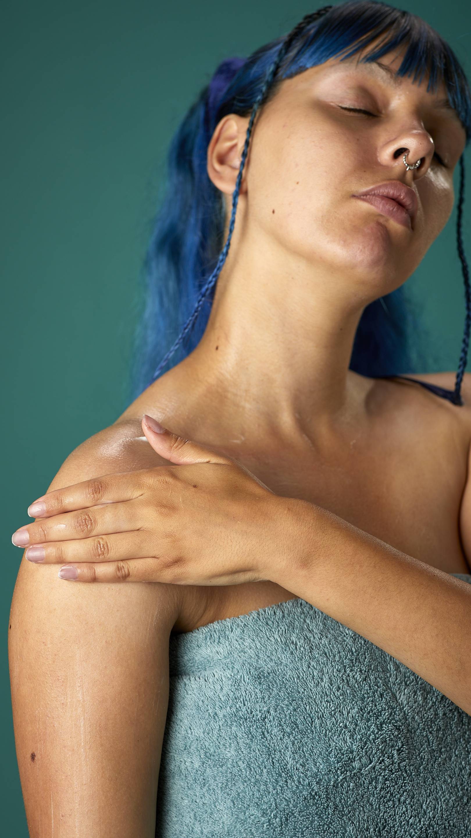 The image shows the model wrapped in a sage-green towel on a teal background. They have blue hair with a full fringe and are gently applying the Coconut body butter to their upper arm.
