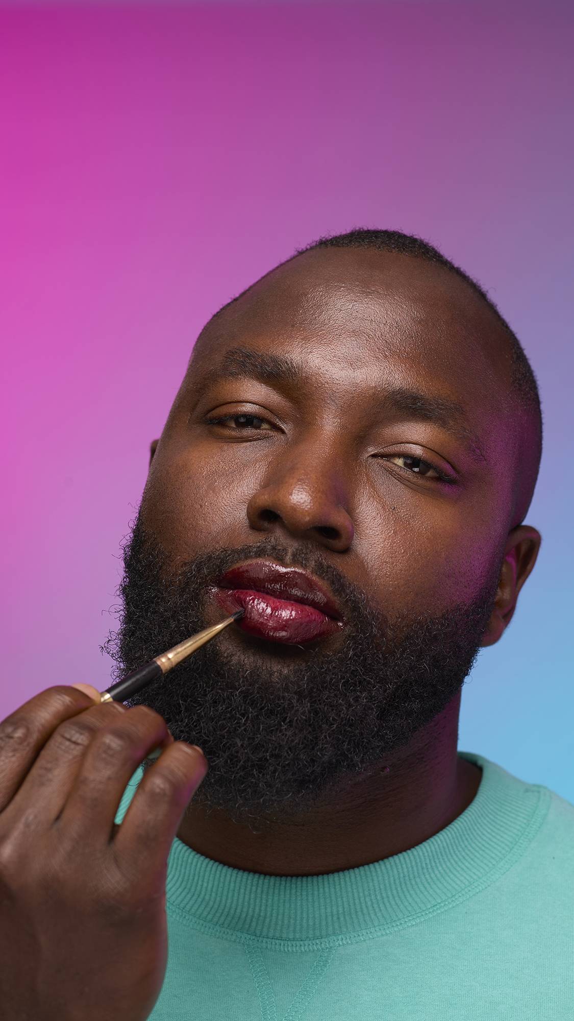 The model is on a pastel pink, blue and purple hue background in a vivid teal jumper. They are using a thin lip brush to apply a juicy coating of grounding, earth-toned tint on their lips created by the Cola lip jelly.