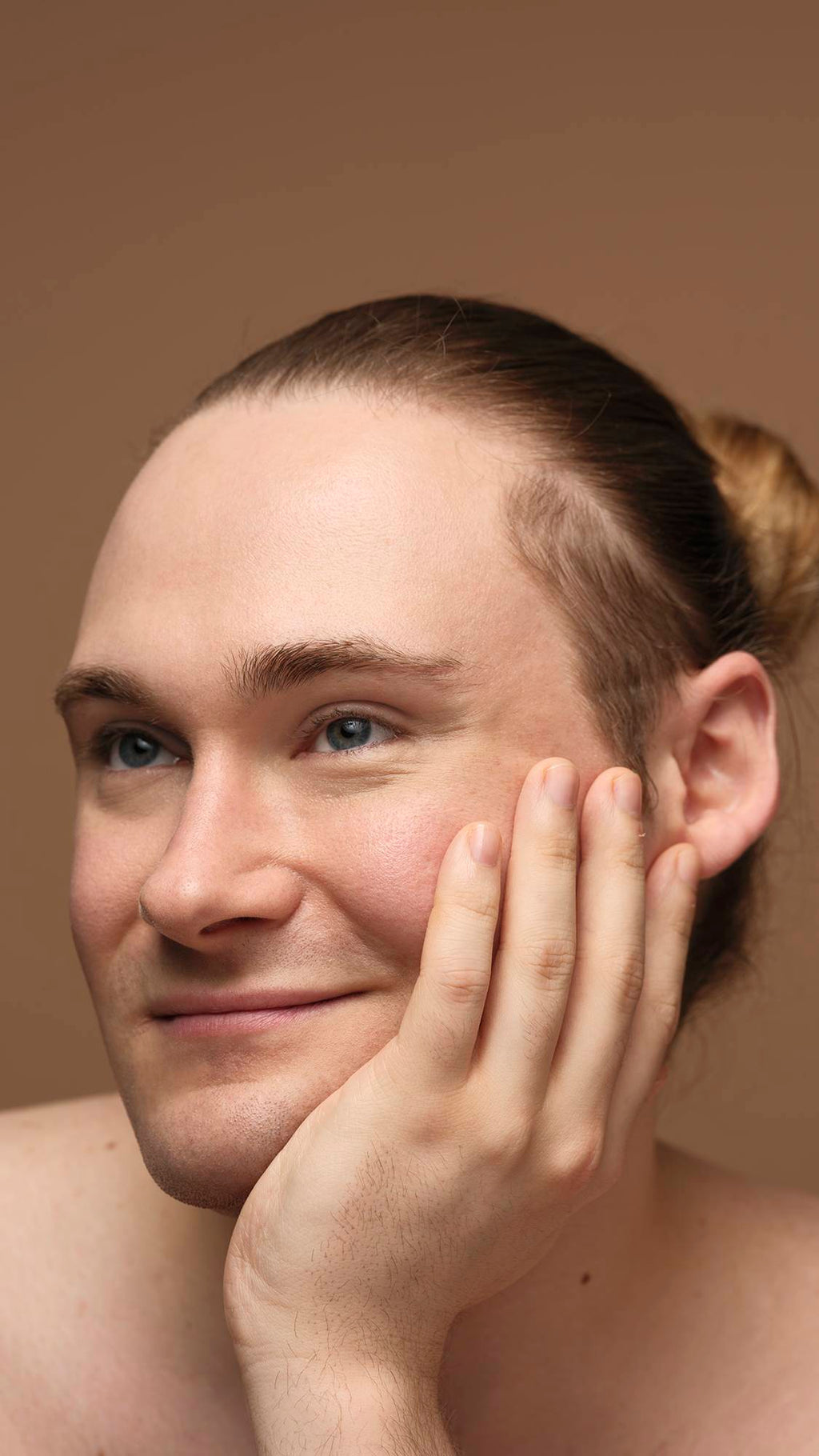 The model is on a warm, earthy-brown background with their head rested upon their hand as they are smiling with glowing, freshly moisturised skin.