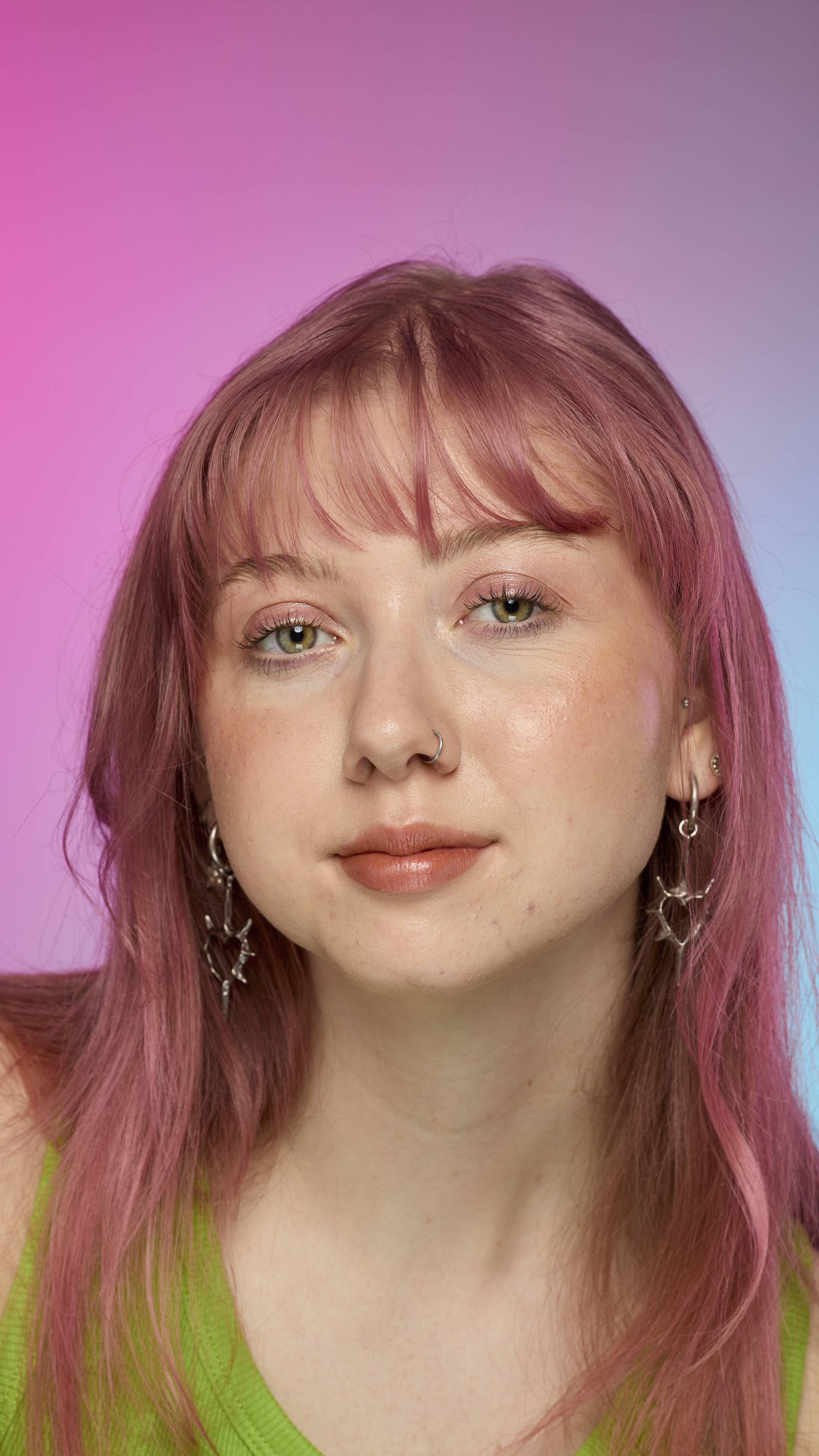 The image shows a fair-skinned model on a pink and blue gradient background. They have pink hair and subtle shimmering lips from using the Crackled Copper lip cream.