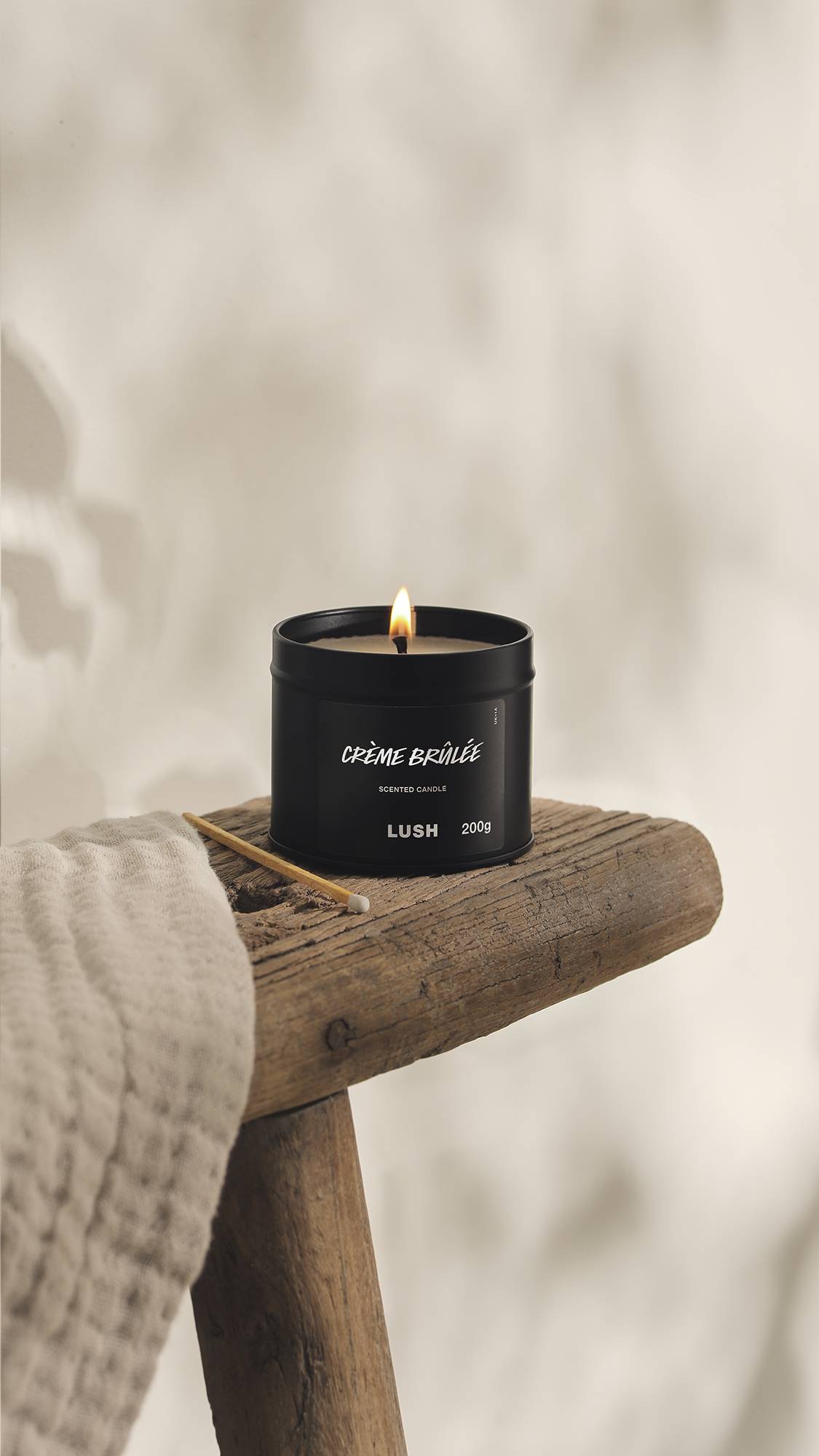 The image shows a blurred, cream background. In the foreground, the Creme Brûlée black tin candle sits with a lit flame on a rustic wooden stool with an unused match beside it and a cream cloth.