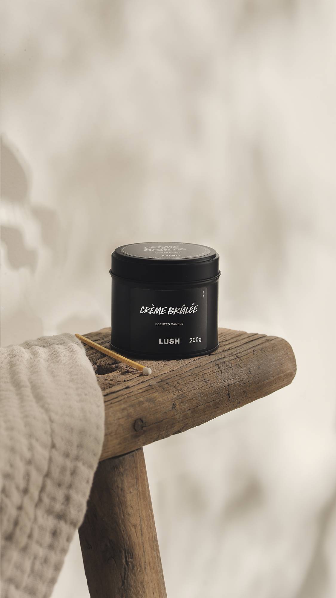 The image shows a blurred, cream background. In the foreground, the Creme Brûlée black tin candle sits on a rustic wooden stool with an unused match beside it and a cream cloth.