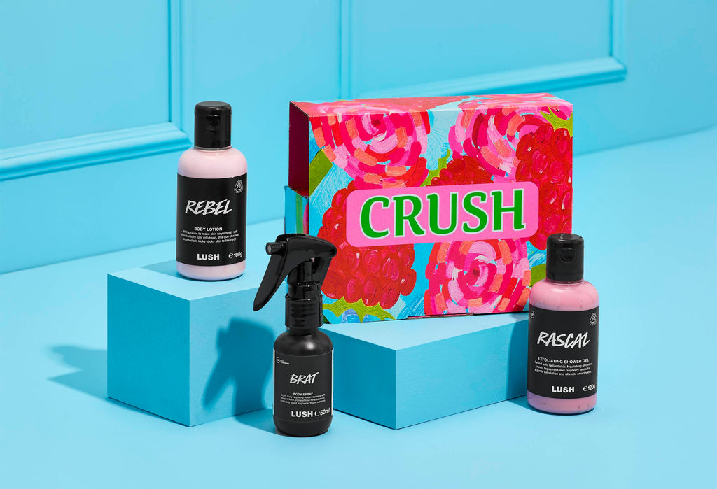 A LUSH Crush gift set displayed on bright blue risers against a matching blue backdrop. The set includes a pink rose-patterned "CRUSH" box, "Rebel" body lotion, "Rascal" shower gel, and "Brat" body spray.