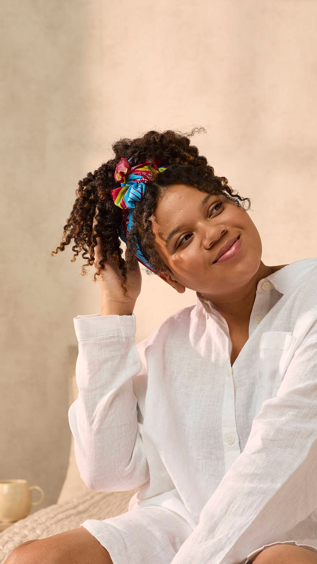 The model is sat on the bed in an oversized white shirt. They have fashioned the Curly Coily gift knot wrap into a trendy head bandana for their thick, curly hair.