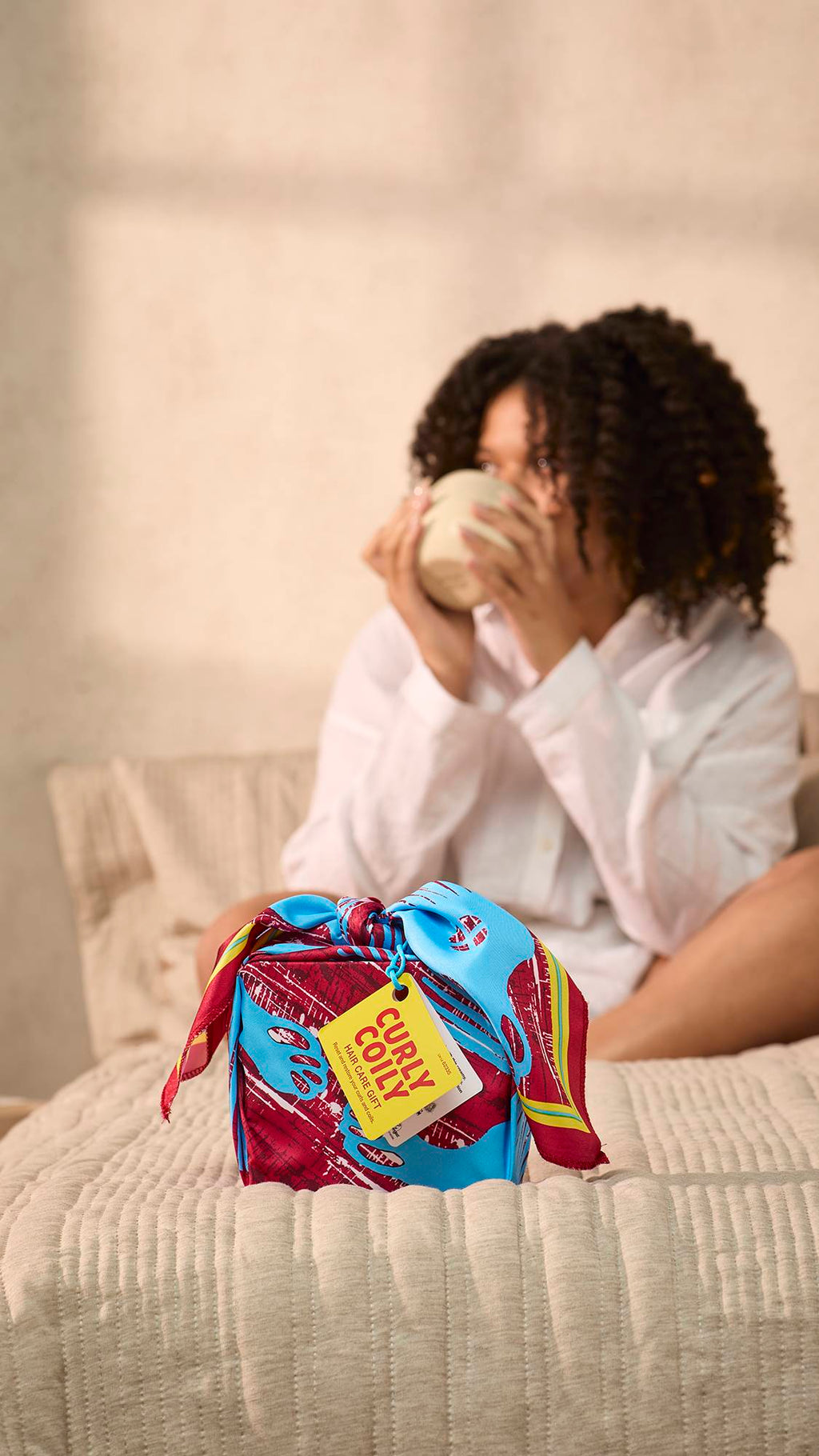 The image shows a light, cream bedroom with the model drinking from a mug in the blurred background as the foreground focuses on the neatly wrapped Curly Coily gift on the end of the bed.
