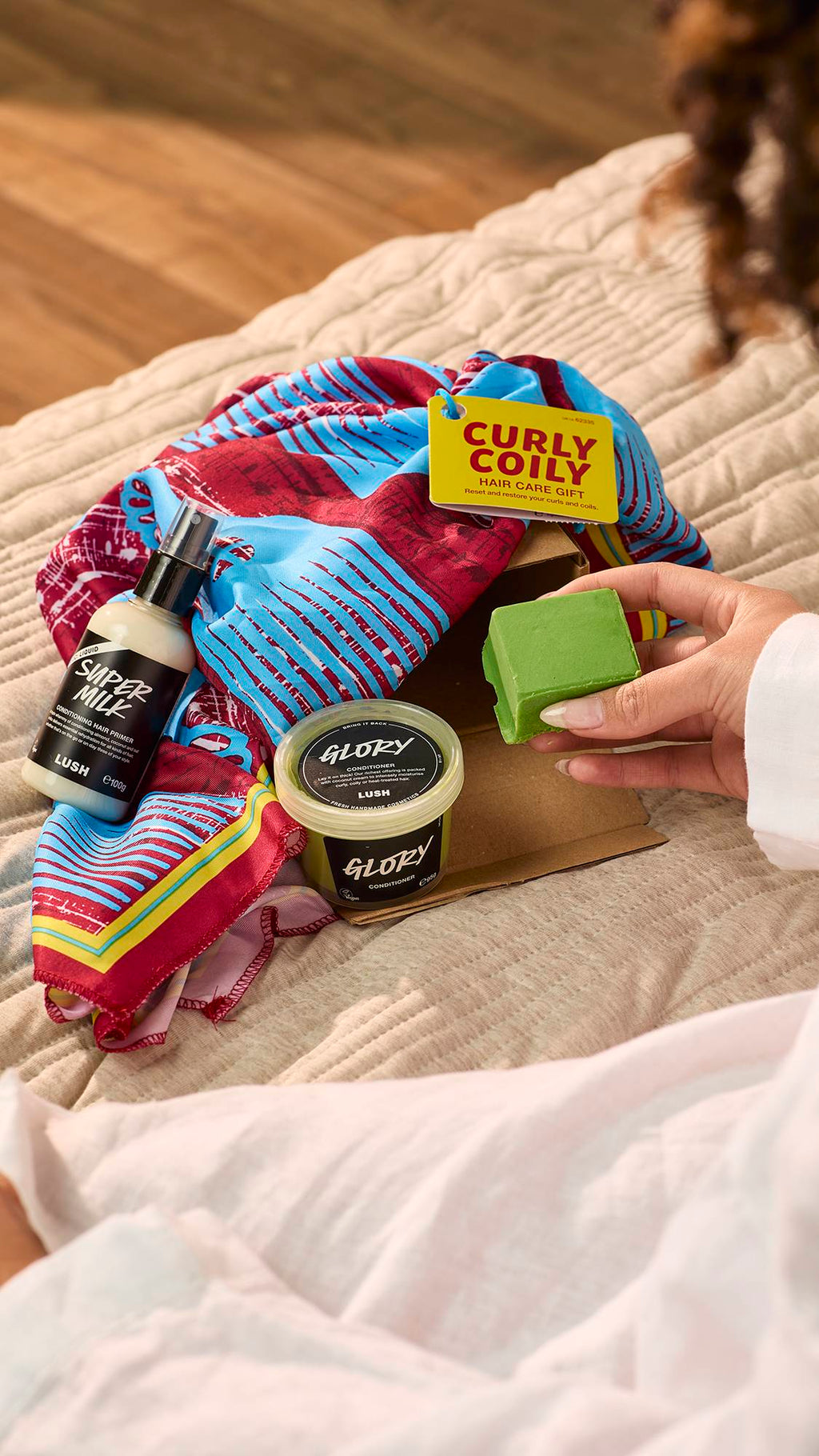 The image shows the Curly Coily gift, having been unpacked, with the three included haircare products on the bed.
