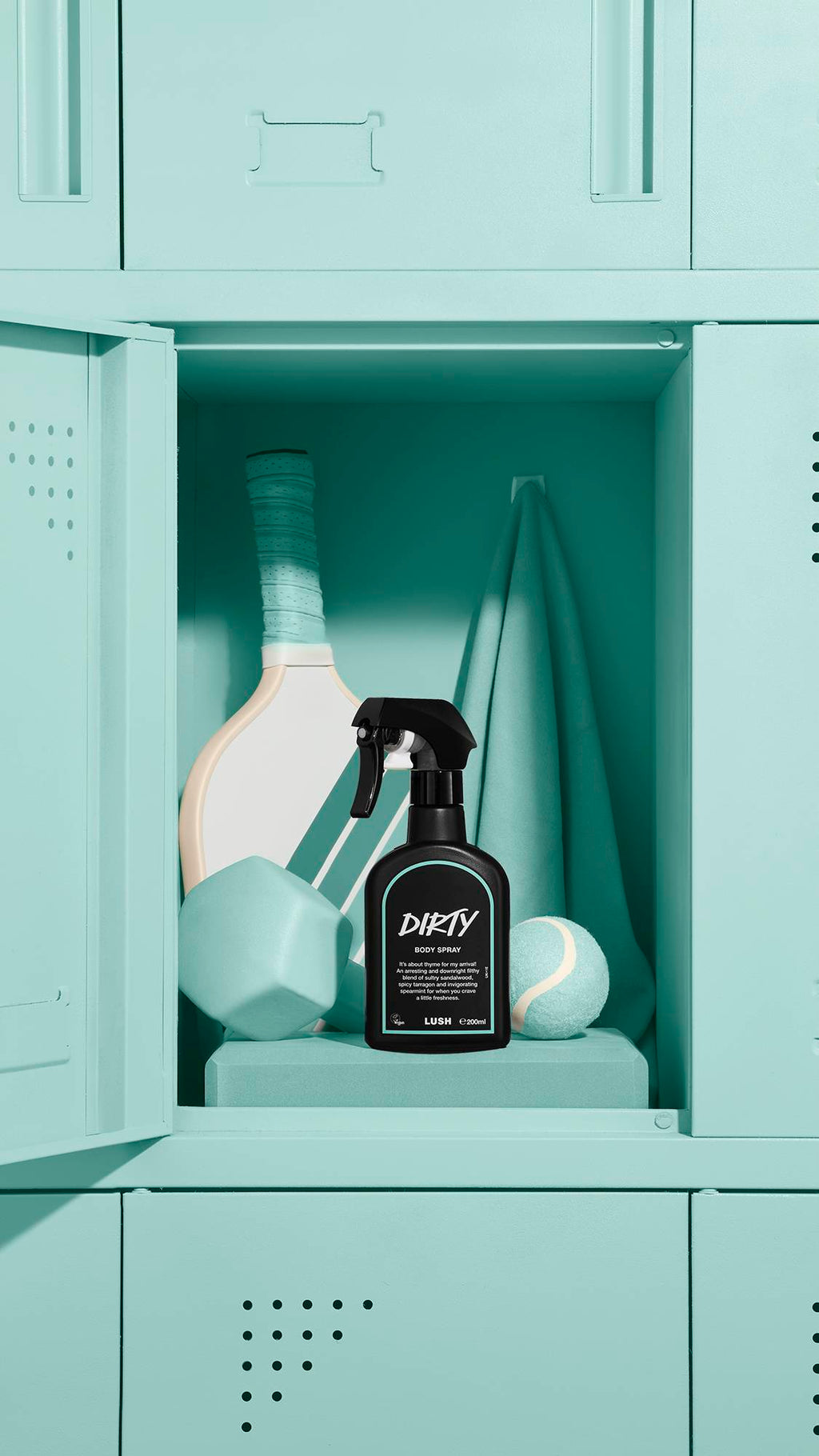 A black bottle of Lush 'Dirty' body spray sits inside an open mint-green locker. Behind the spray are a matching green paddle, towel, tennis ball, and geometric foam blocks, creating a cohesive, sporty aesthetic.