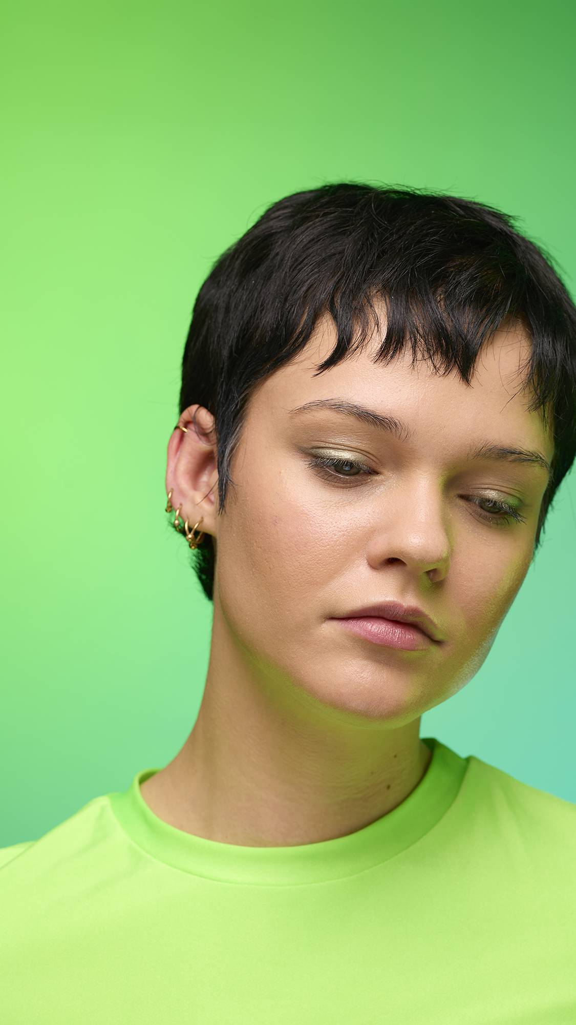 The image shows a close-up of the model's face on a pale blue-to-green gradient background. The model's make-up is done with the focal point being a subtle, gunmetal-brown, shimmering eyeshadow made by the Eclipse Glitter Crumble.