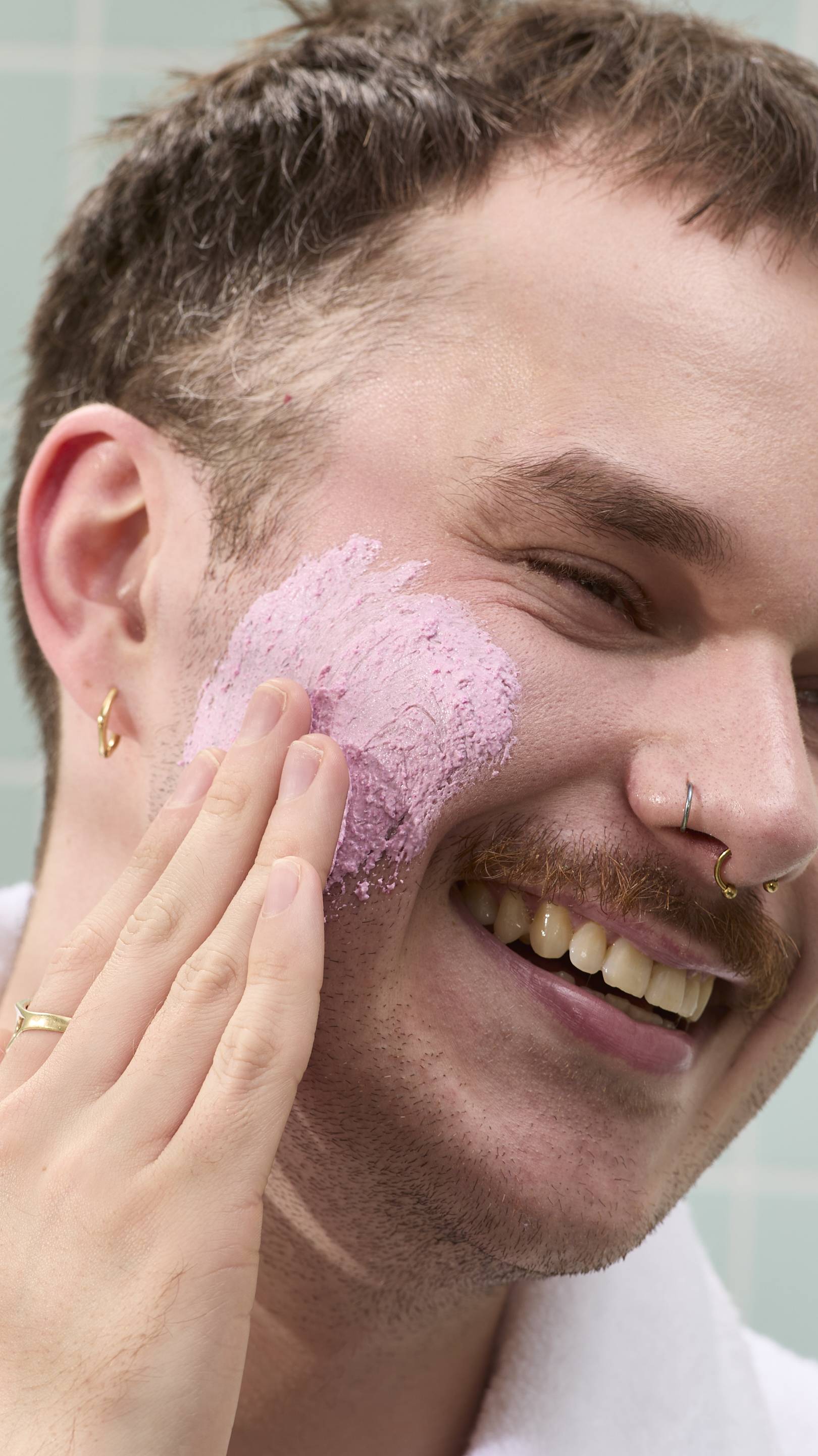 A model with a moustache and nose piercings smiles while applying a thick, grainy pink "Family Face Time" face mask to their cheek. They are wearing a white dressing gown against a background of pale green tiles.