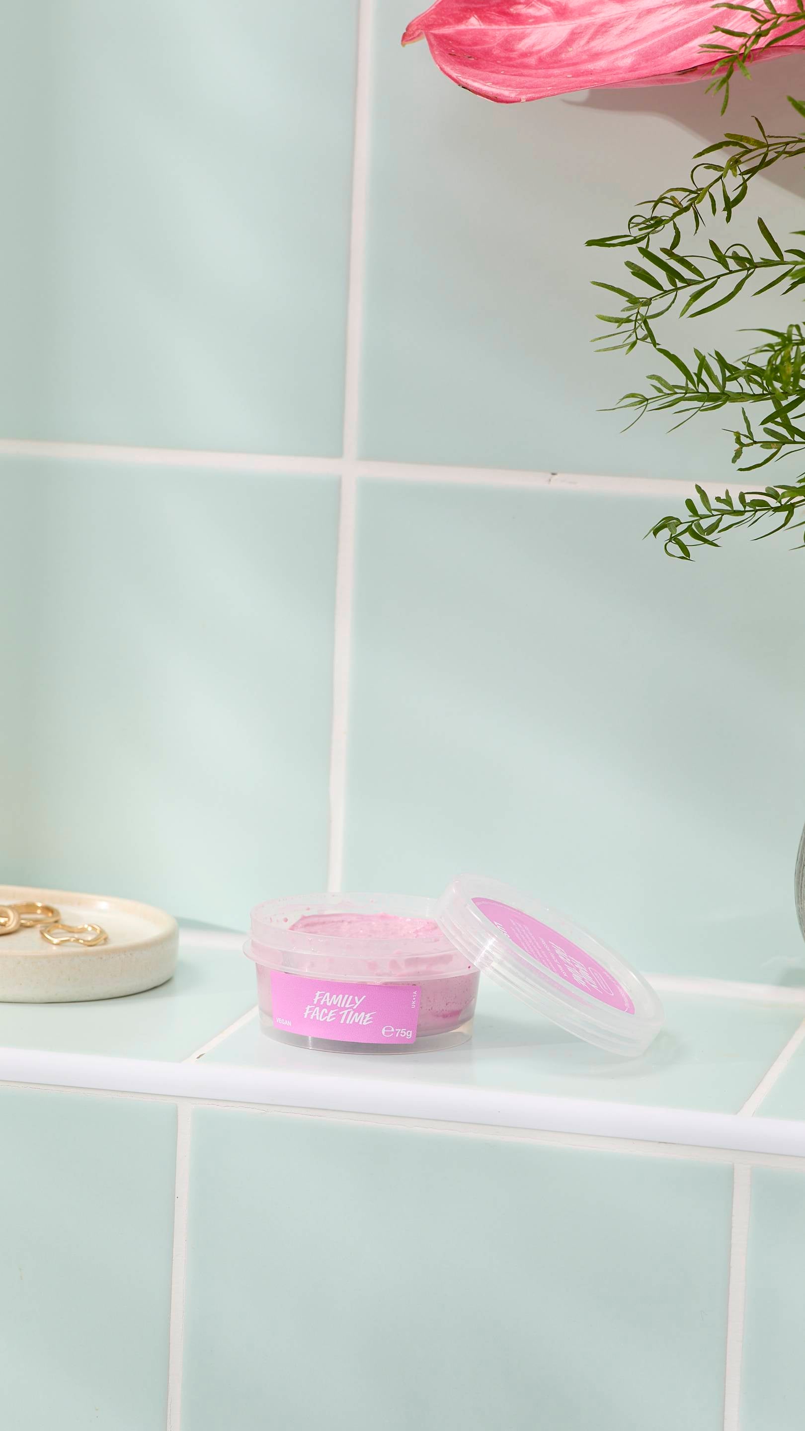 An open, clear plastic pot of 'FAMILY FACE TIME' fresh face mask sits on a pale green tiled ledge. The pot contains a thick, grainy pink face mask, with some residue on the lid. A small dish with gold rings and a pink flower are nearby.