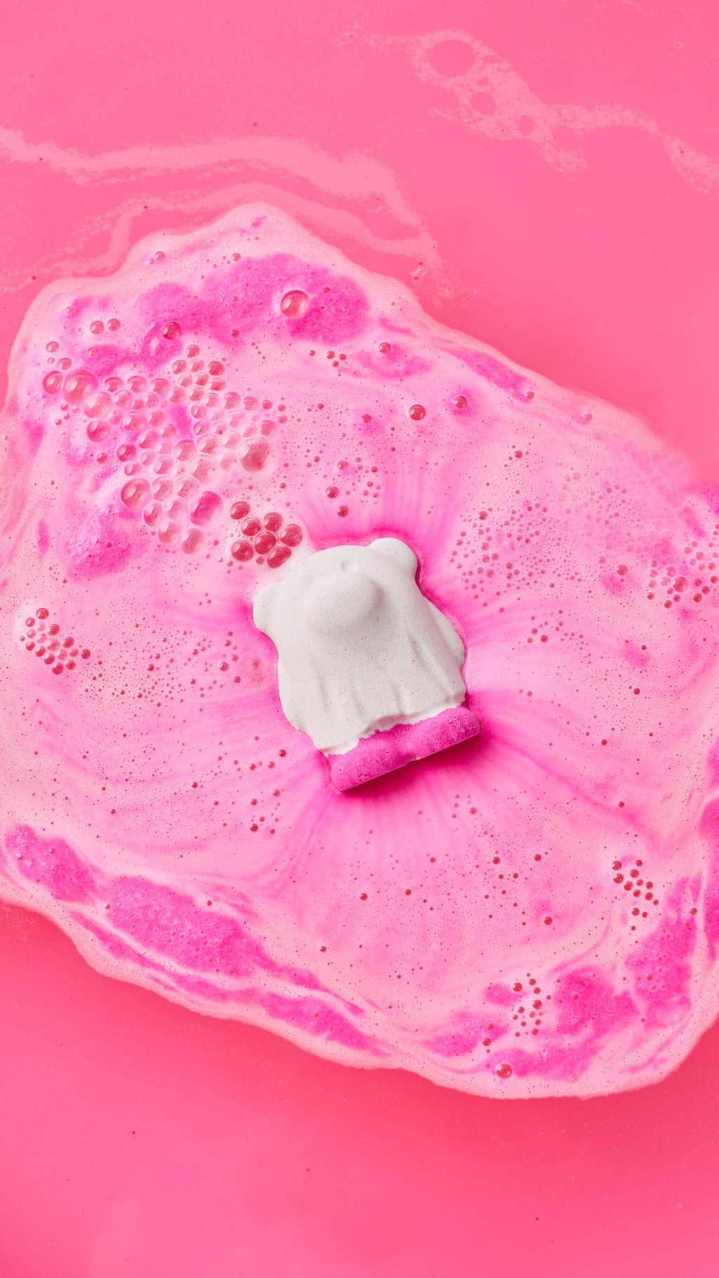 A white ghost bear bath bomb fizzes and dissolves in bright pink bathwater, floating in a pool of vibrant pink and white foam and bubbles.