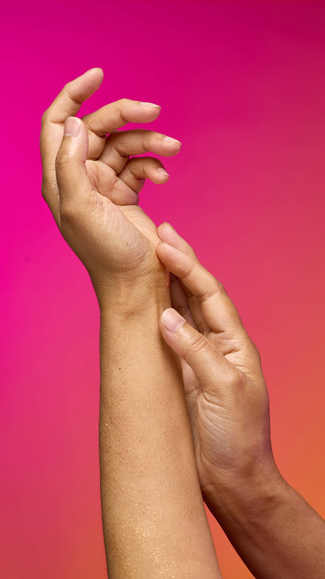The image shows a model's forearm extended from the bottom as the skin appears moisturised with a subtle, delicate shimmer over the skin after having used the Golden Egg Glitter Mist spray product on a pink-to-orange gradient background.