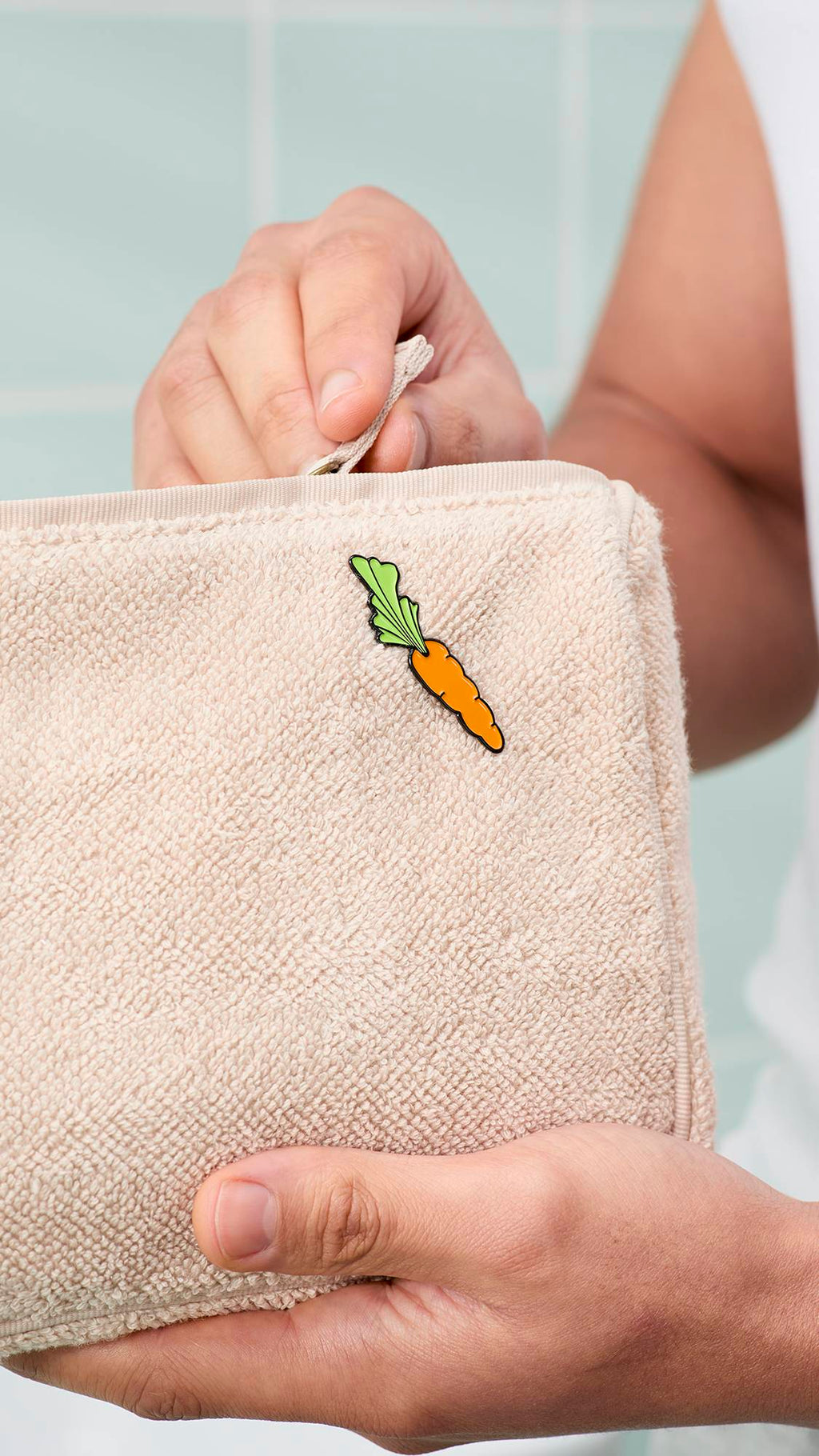 A person's hands hold a beige, textured towelling wash bag featuring a small enamel carrot pin from the "Happy Bunny" greetings card. One hand is shown pulling the zip closed against a background of mint-green bathroom tiles.