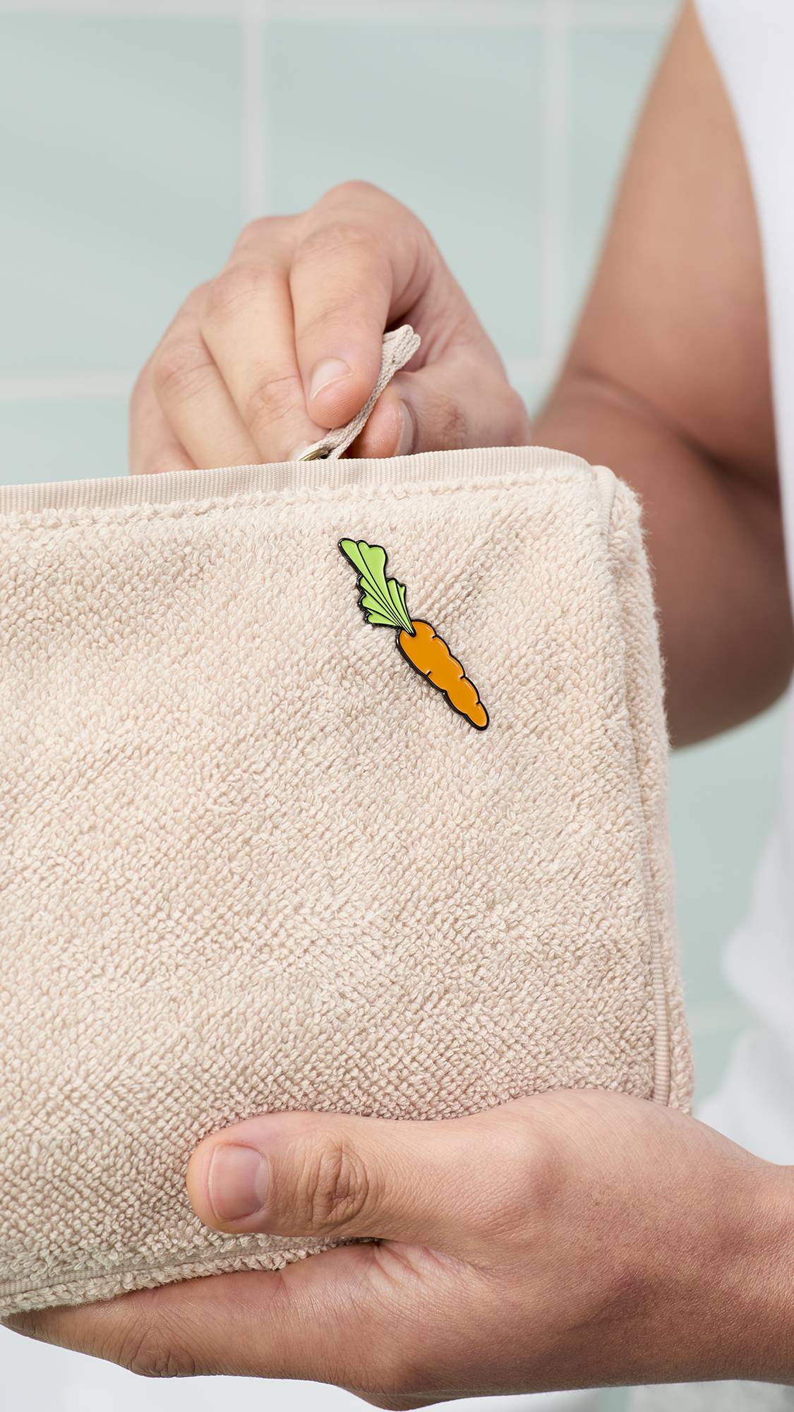 A person's hands hold a beige, textured towelling wash bag featuring a small enamel carrot pin from the "Happy Bunny" greetings card. One hand is shown pulling the zip closed against a background of mint-green bathroom tiles.