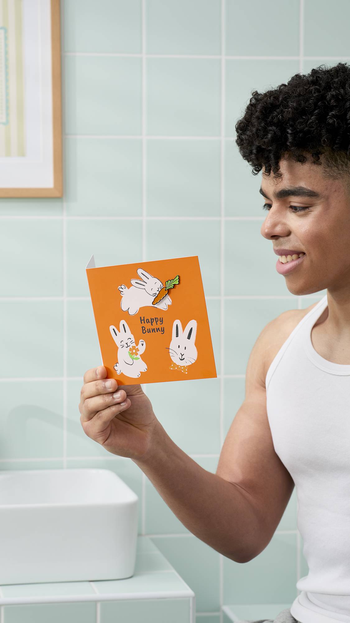 A model with dark, curly hair and a white vest smiles while holding an orange "Happy Bunny" greeting card. They are in a bathroom with pale mint-green tiles, a white basin, and a framed picture on the wall.