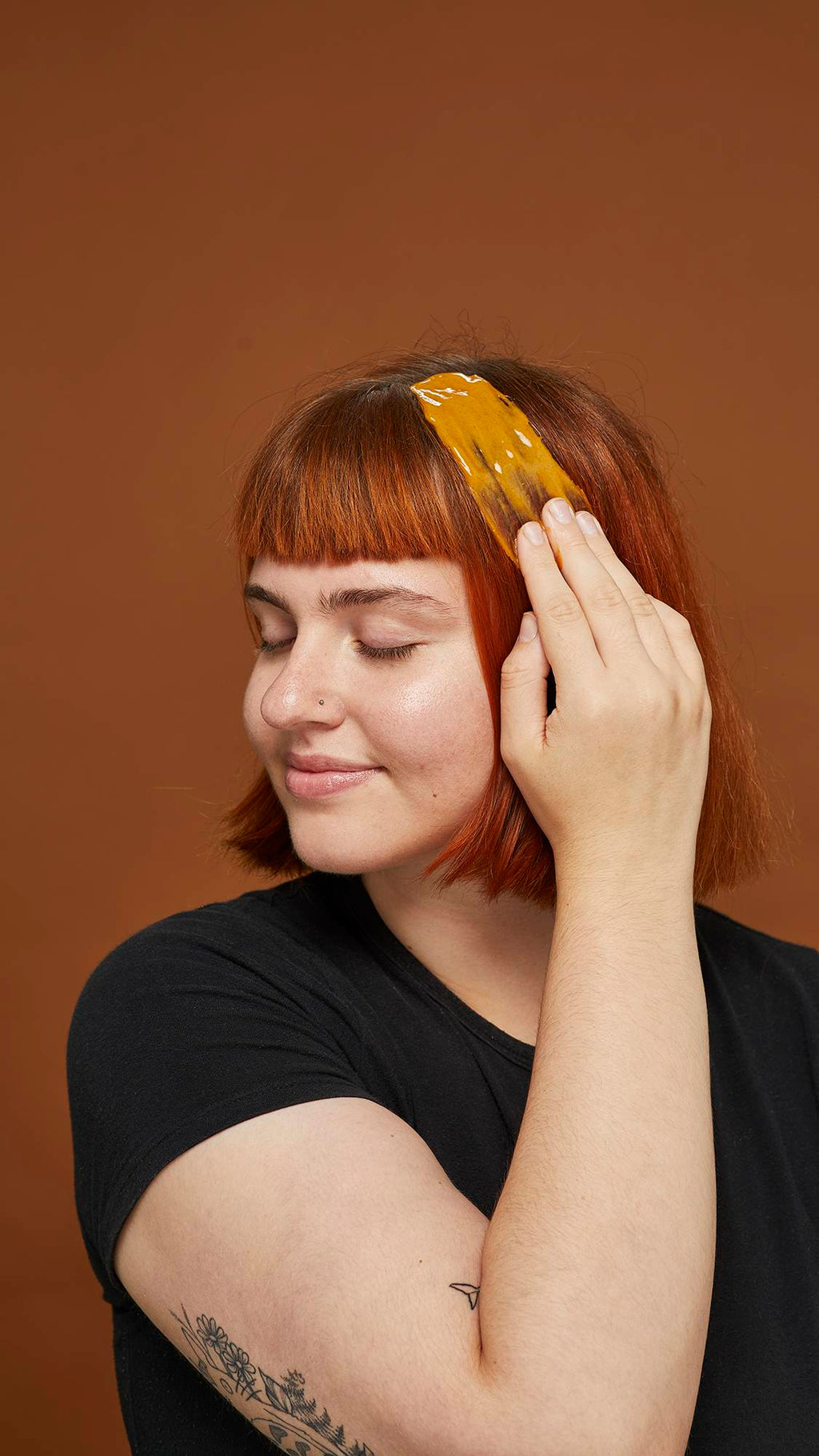 Image shows model slicking on the cooled-down hot oil treatment over auburn-coloured hair on an earth-toned background.