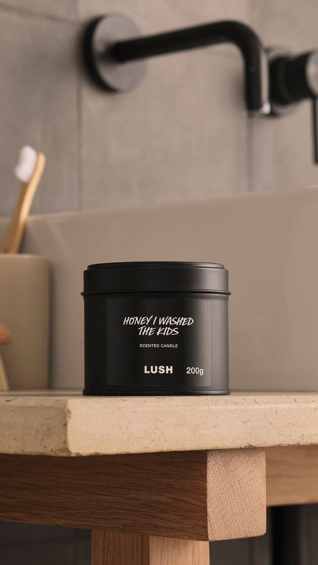 The image shows a blurred, grey background with a black tap and bamboo toothbrush. In the foreground, the Honey I Washed The Kids black tin candle with the lid sits atop a wooden, stone-topped counter.