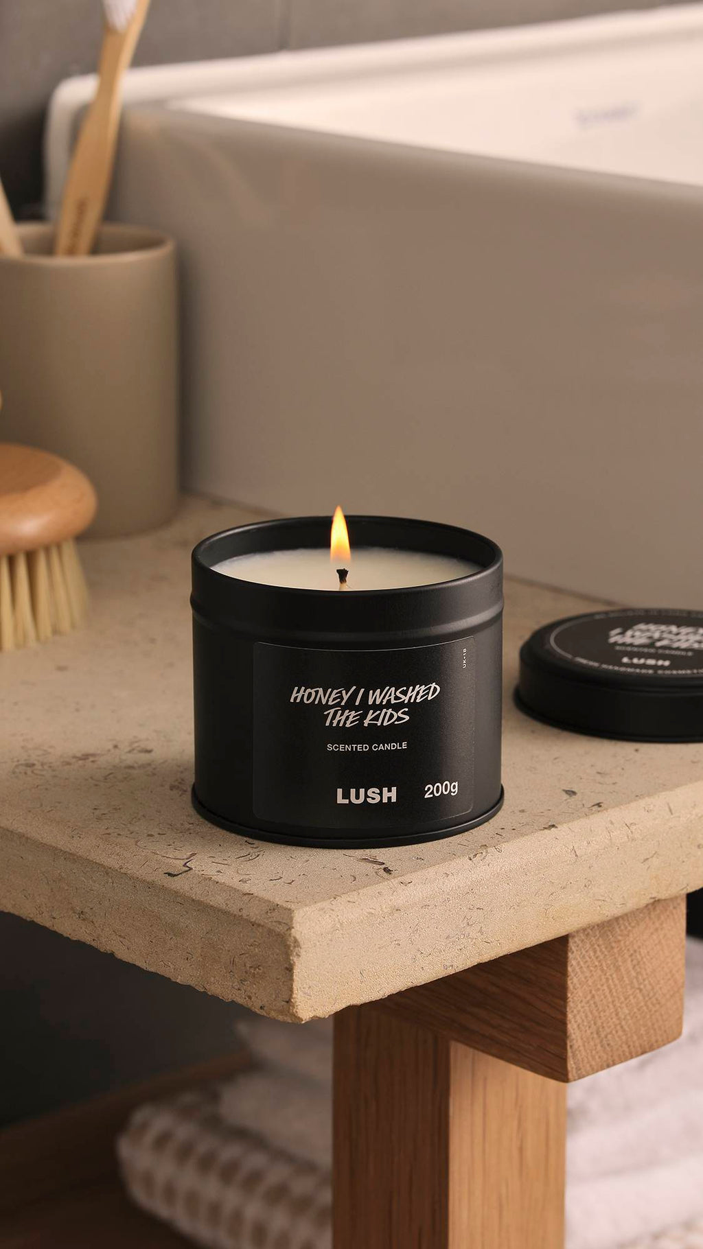 The image shows a blurred background with a white, square bath and bamboo toothbrush. In the foreground, the lit Honey I Washed The Kids black tin candle sits atop a wooden, stone-topped counter.