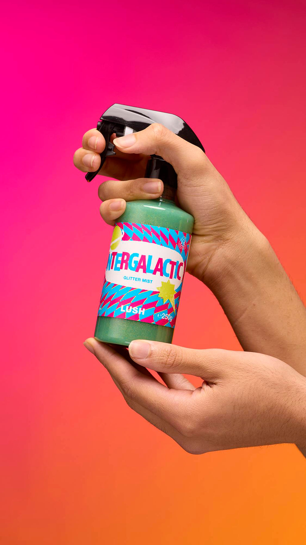 An arm is reaching from the right-hand side. The hand holds a transparent, cylindrical plastic spray bottle with teal, shimmering liquid and a black top. A colourful label reads "Intergalactic Glitter Mist" on a pink-to-orange gradient background.