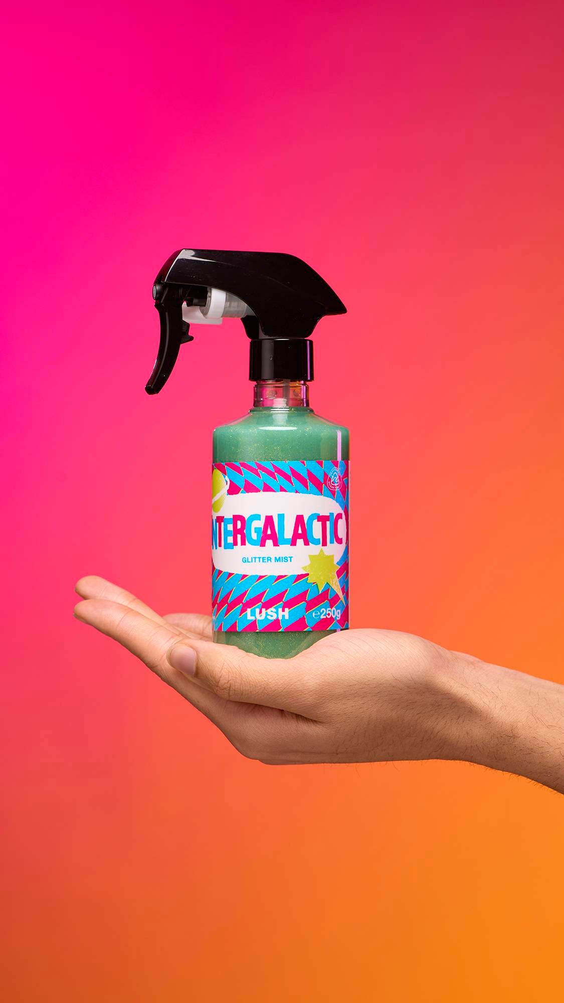 An arm is reaching from the right-hand side. The hand holds a transparent, cylindrical plastic spray bottle with teal, shimmering liquid and a black top. A colourful label reads "Intergalactic Glitter Mist" on a pink-to-orange gradient background.