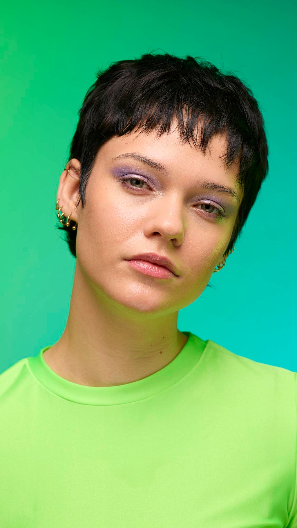 The image shows a close-up of the model's face on a pale blue-to-green gradient background. The model's make-up is done with the focal point being a delicate, purple, shimmering eyeshadow made by the Little Sparkler Glitter Crumble.