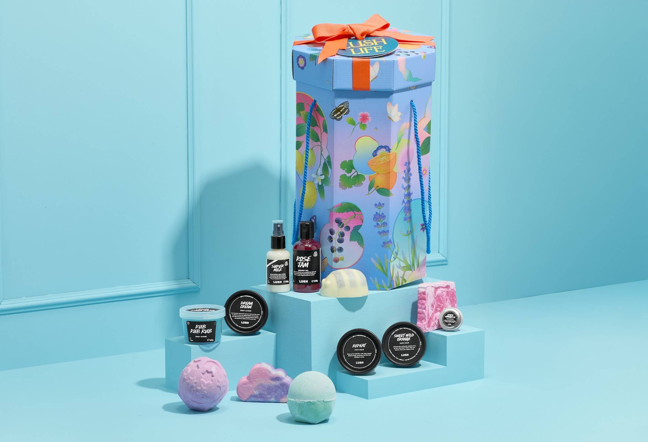 The image shows a tall, hexagonal 'Lush Life' gift box with colourful illustrations, sitting on a blue plinth. A selection of 12 products are arranged on steps around it, against a light blue panelled wall.