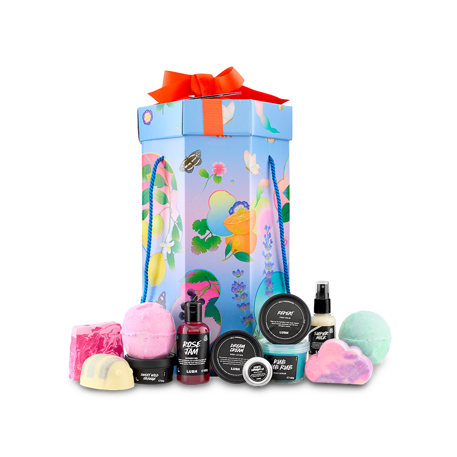 The image shows the 'Lush Life' gift set. A tall, hexagonal gift box with colourful illustrations sits behind a selection of 12 products, including bath bombs, soap, and potted products.