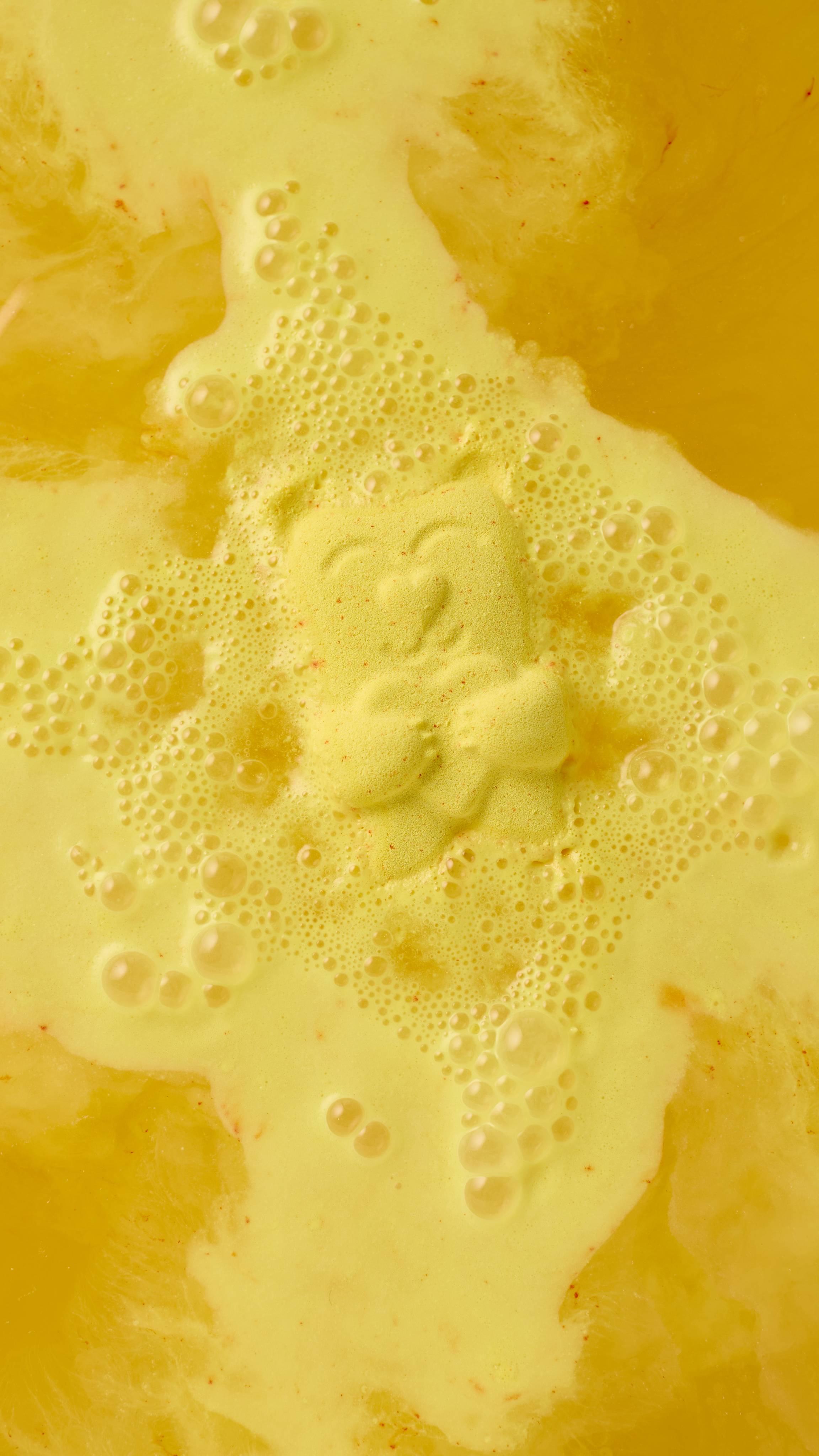 A close-up, top-down view of a yellow "Mama Bear" bath bomb dissolving in water. The bear fizzes in the centre, creating a bright yellow, foamy lather and bubbles that spread across the surface of the golden-yellow bathwater.